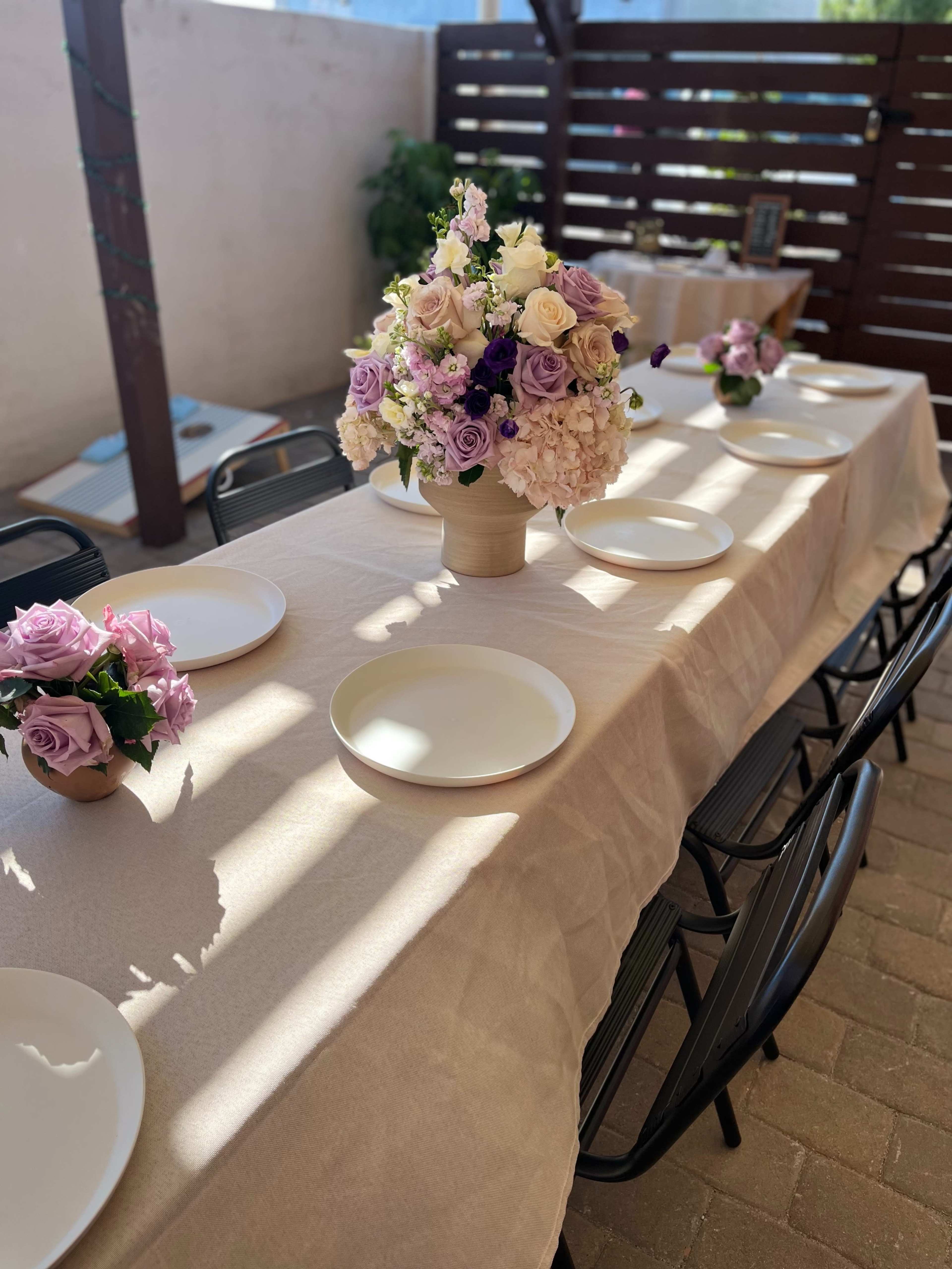 A long table is set with white plates and floral centerpieces in a well-lit outdoor space.