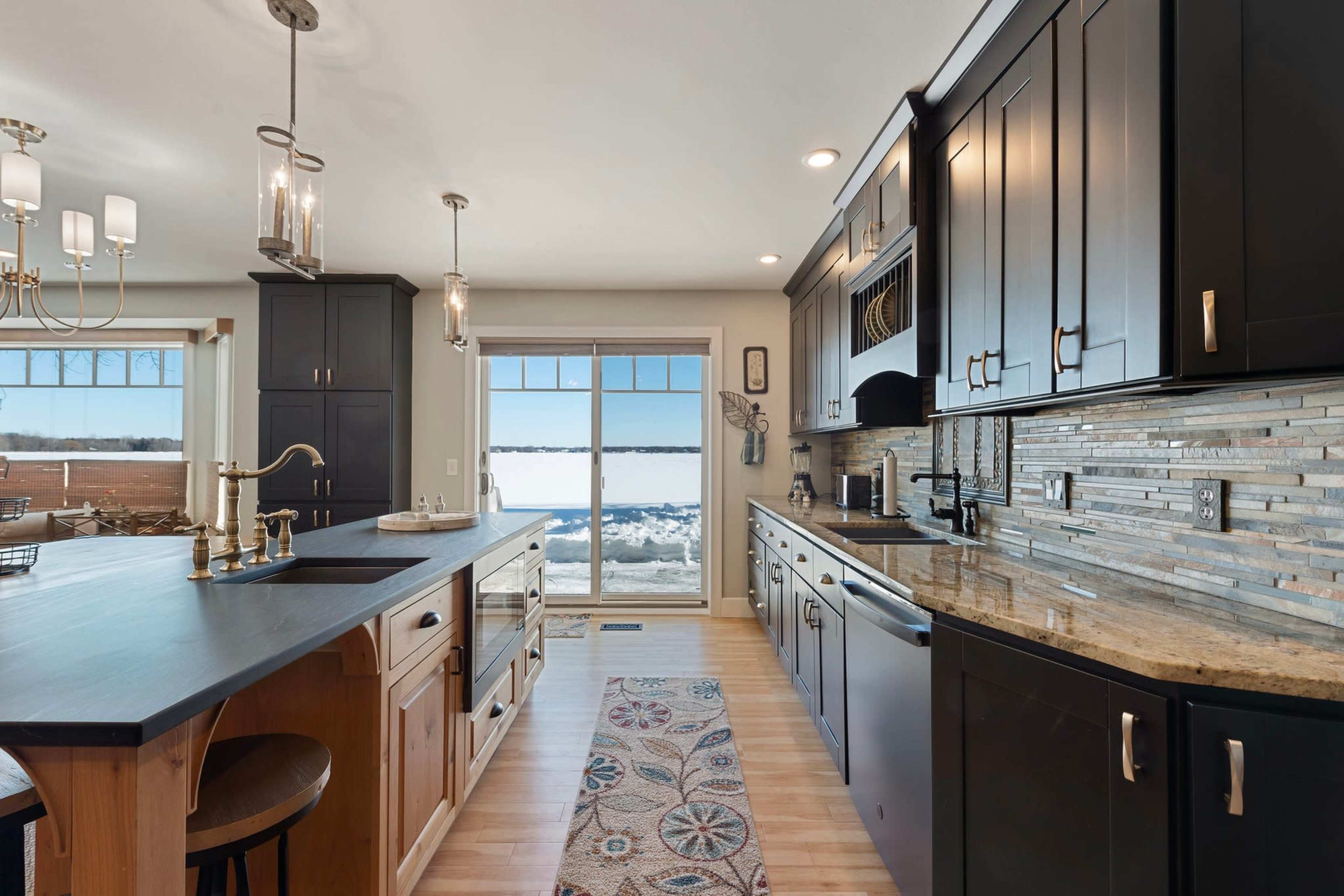 A modern kitchen features dark cabinetry, a large island with bar stools, and a view of the snowy landscape through a wide window.
