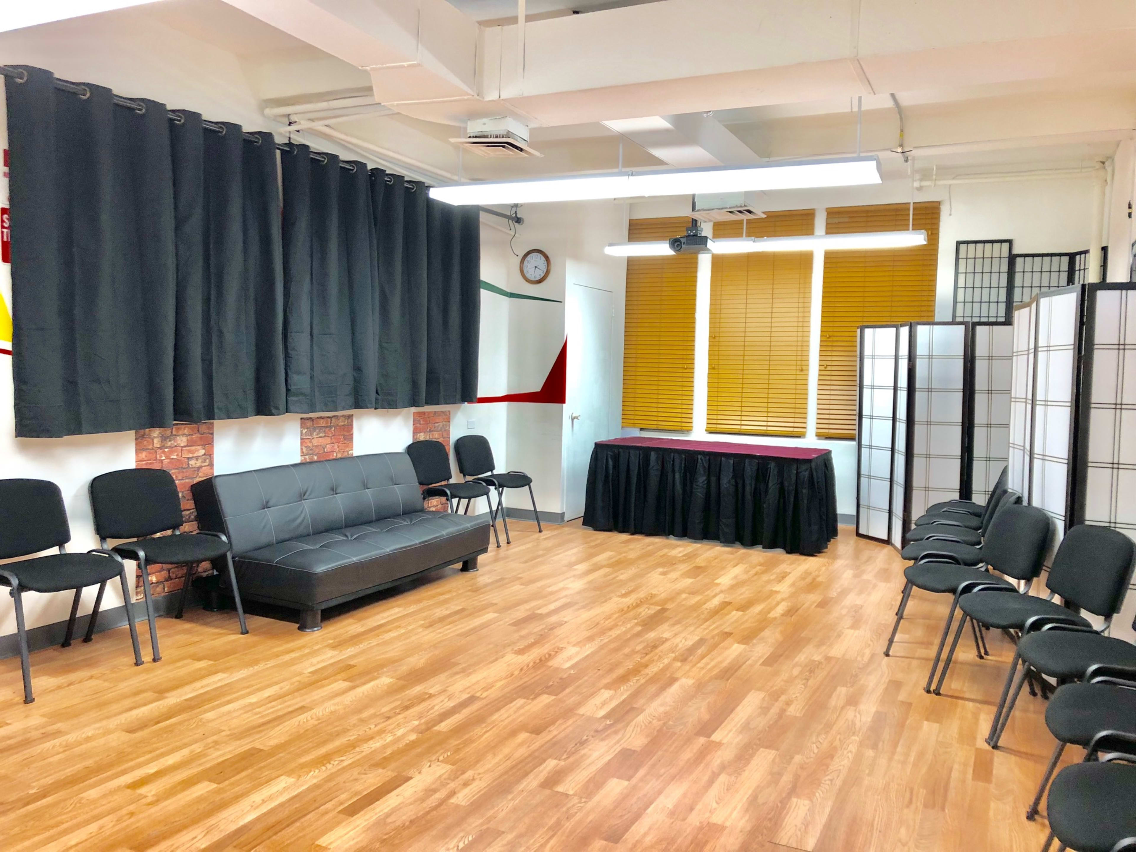 The room features a dark couch and several black chairs arranged in a semi-circle, with a table covered in a cloth at one end and large curtains along one wall.