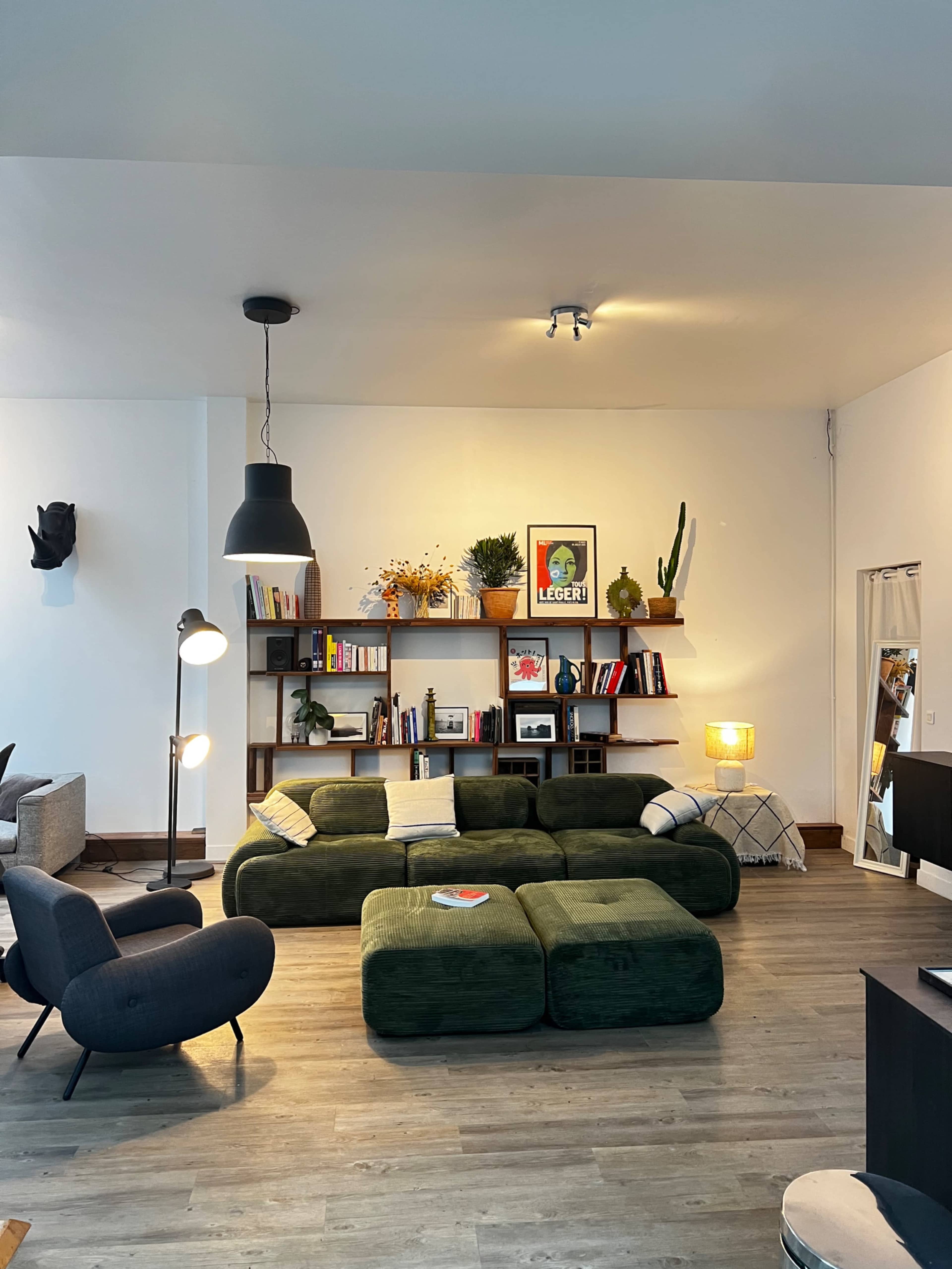 The image shows a modern living room with a green modular sofa, bookshelves filled with books and decor, and various lighting elements.