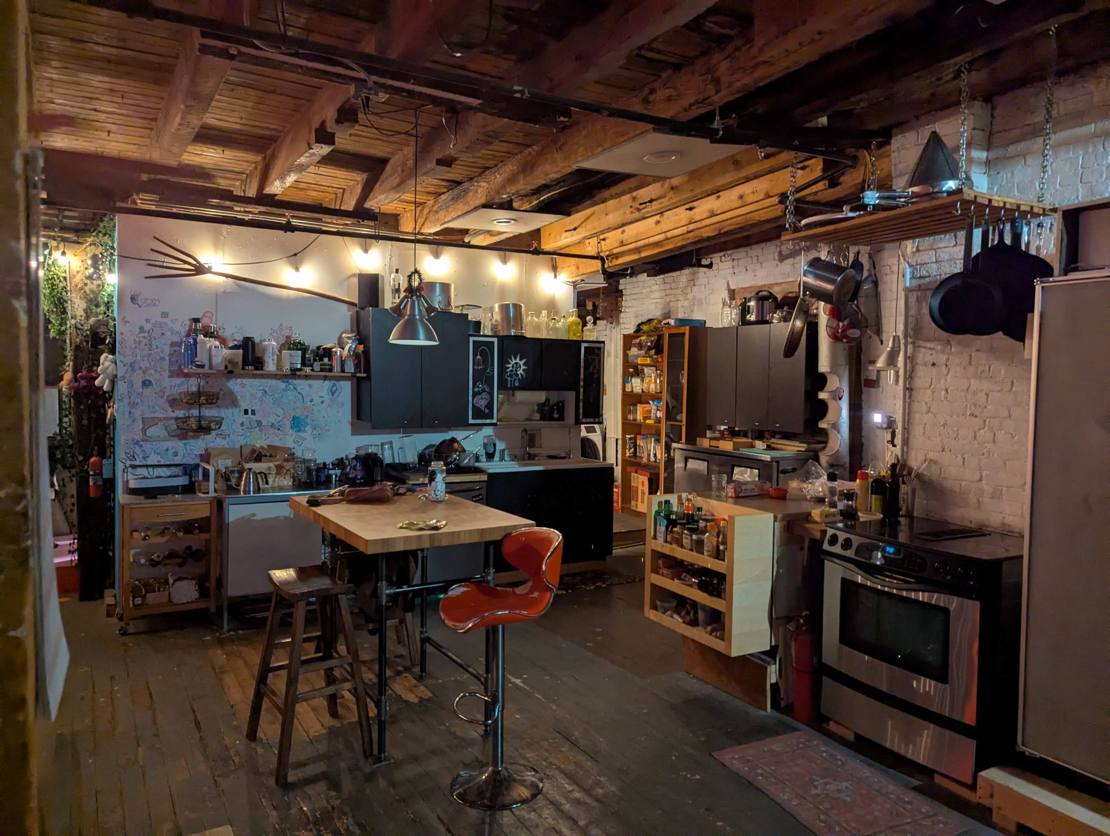 The image shows a rustic kitchen with wooden beams, a central table, black cabinets, and various cooking utensils hanging from the ceiling.