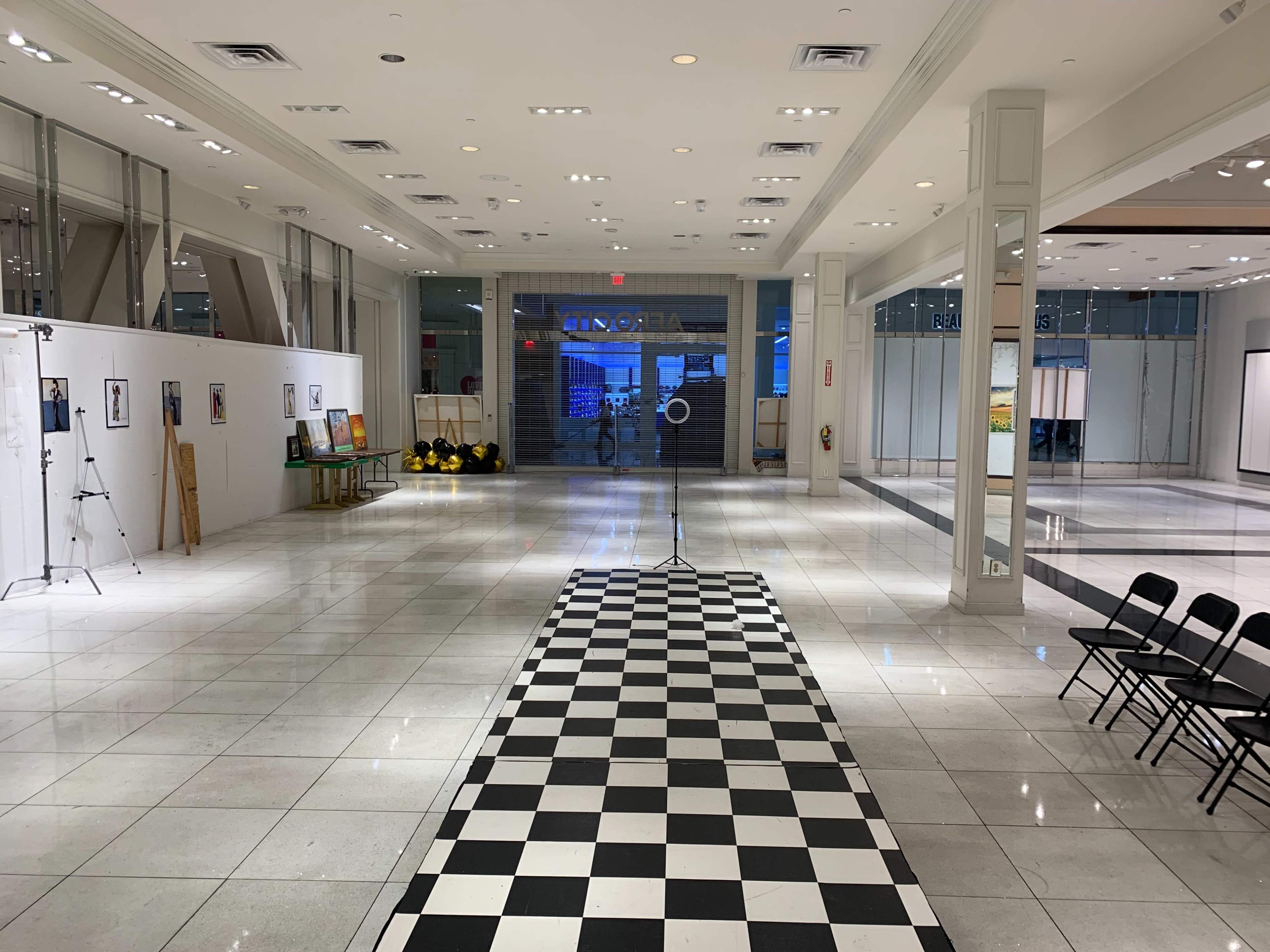 An empty gallery space with a checkered floor, white walls displaying artwork, and rows of black chairs set up in front of a closed entrance.
