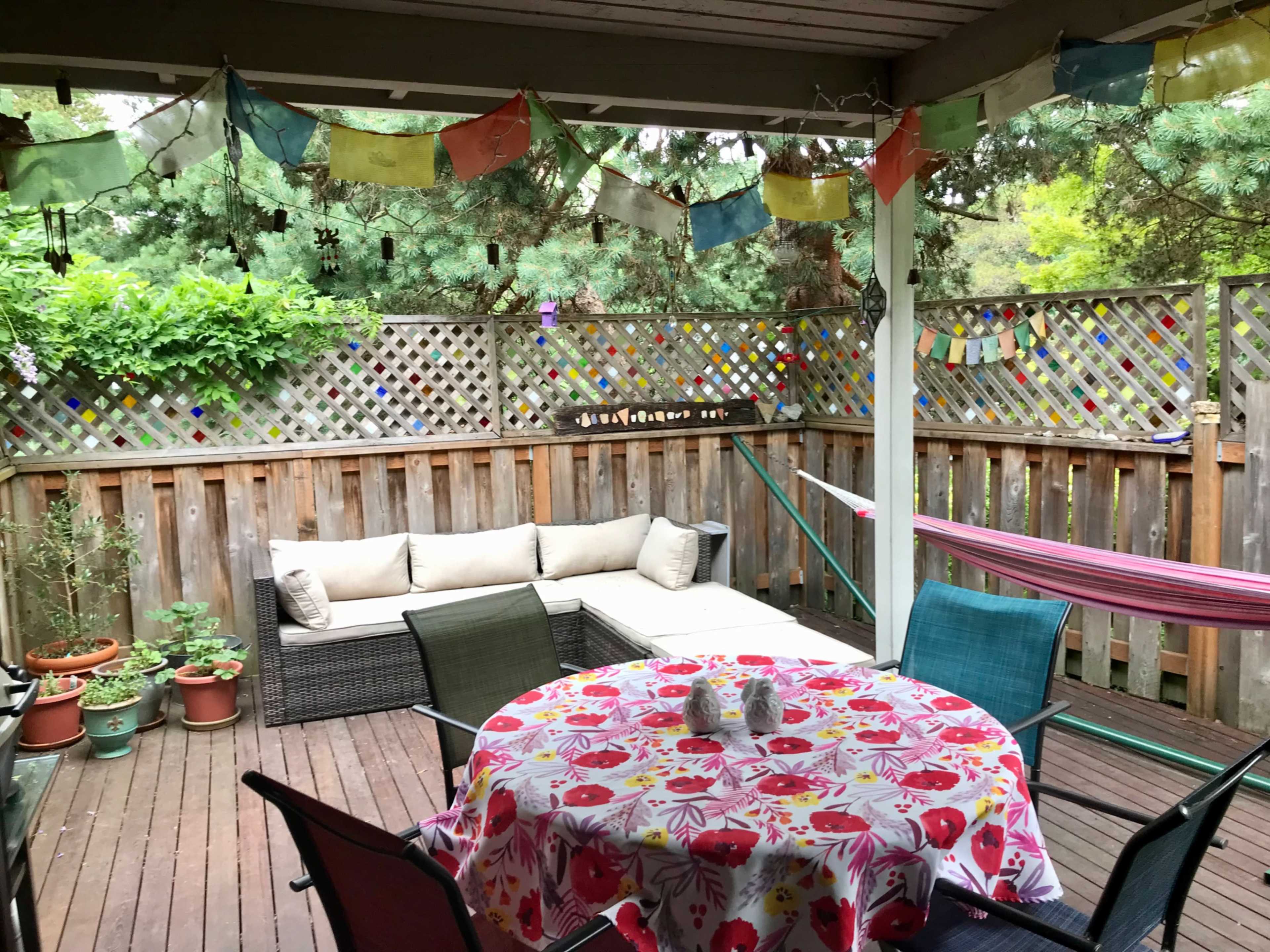 A cozy outdoor patio area features a round table with a floral tablecloth, surrounded by chairs, and decorative prayer flags hanging overhead amidst potted plants.
