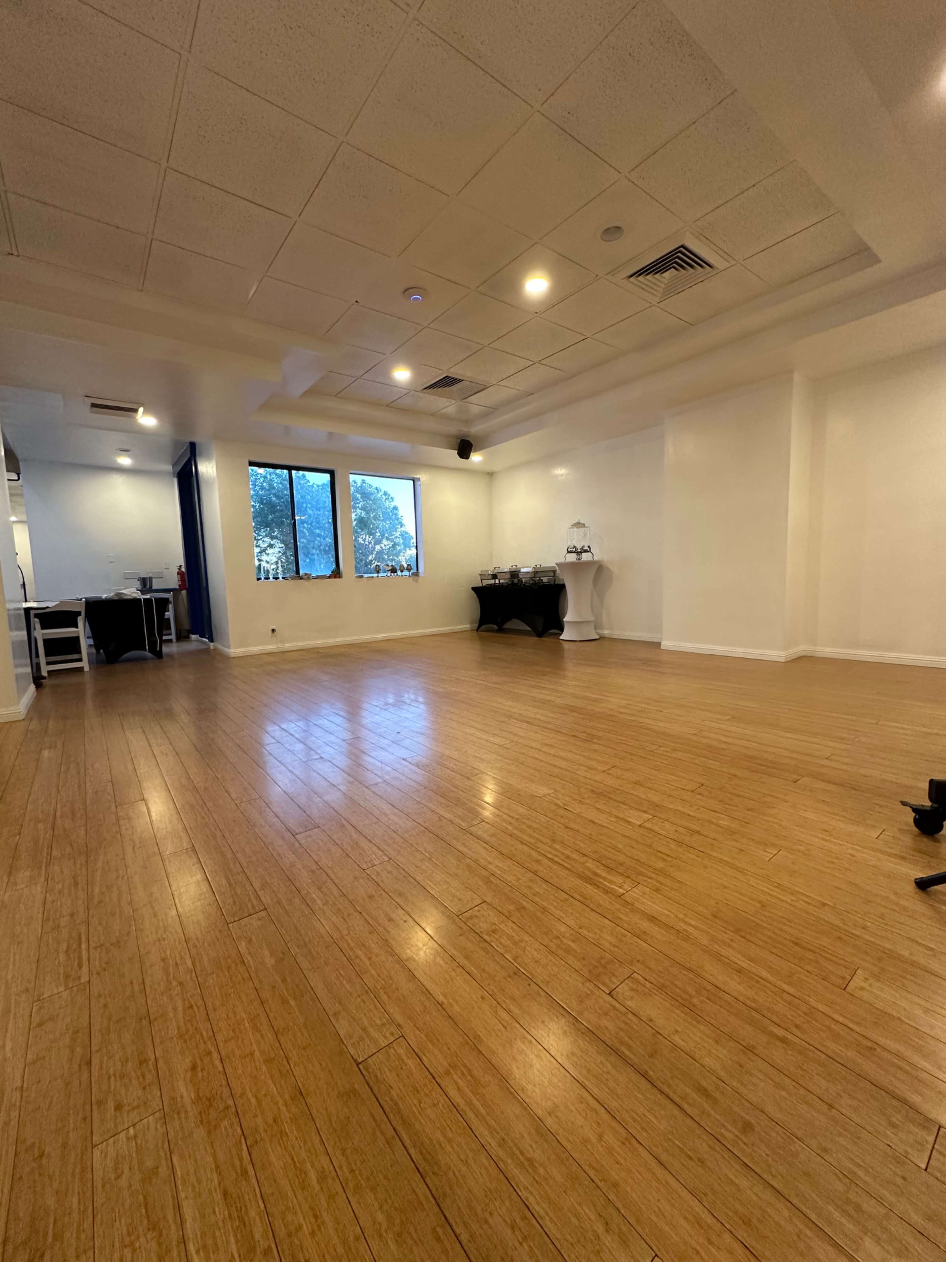 The image shows a spacious, empty room with wooden flooring, large windows letting in natural light, and minimal furnishings.