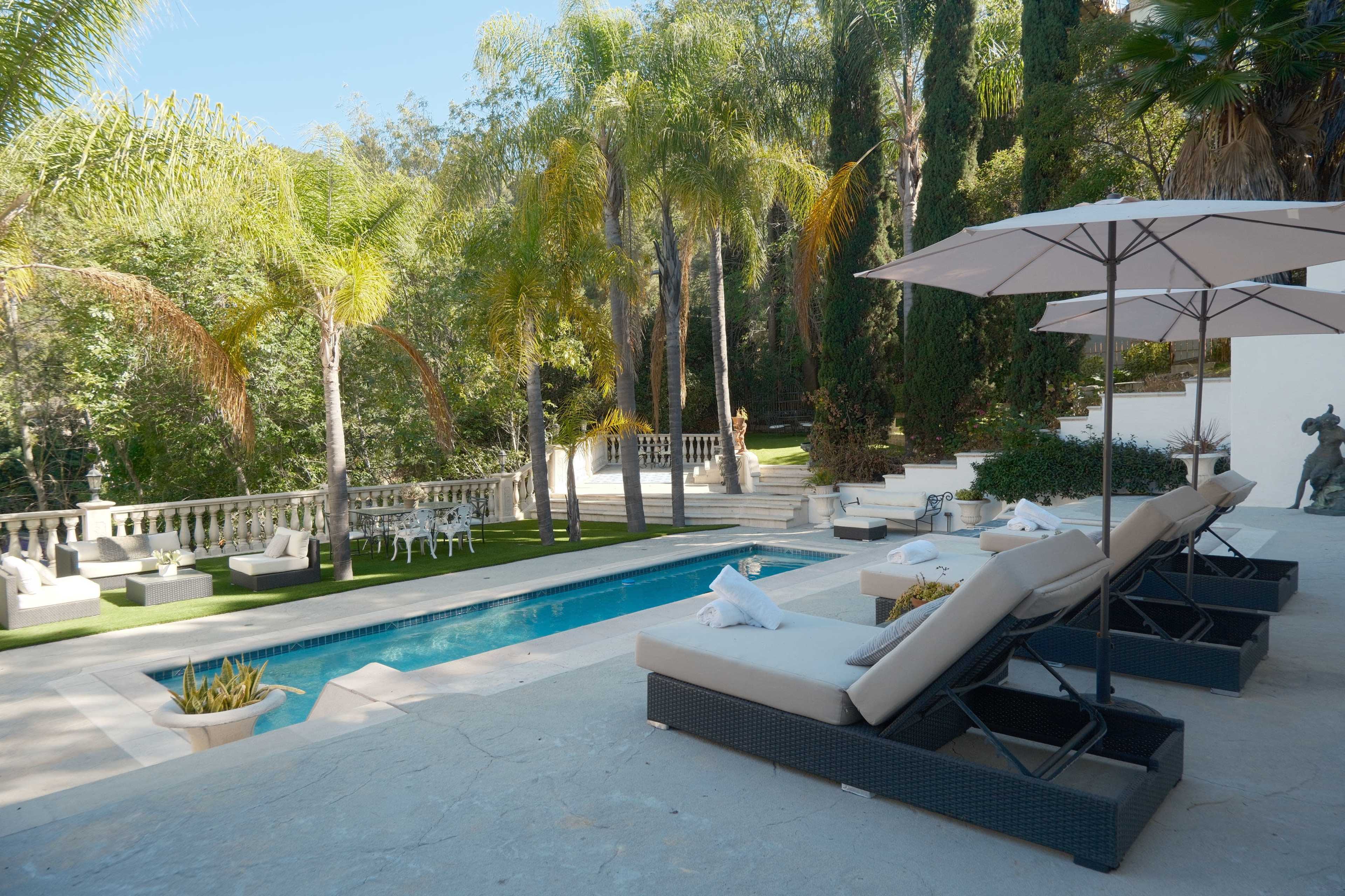 The image shows a tranquil outdoor area featuring a swimming pool bordered by lounge chairs and palm trees, with an adjacent patio and landscaped greenery.
