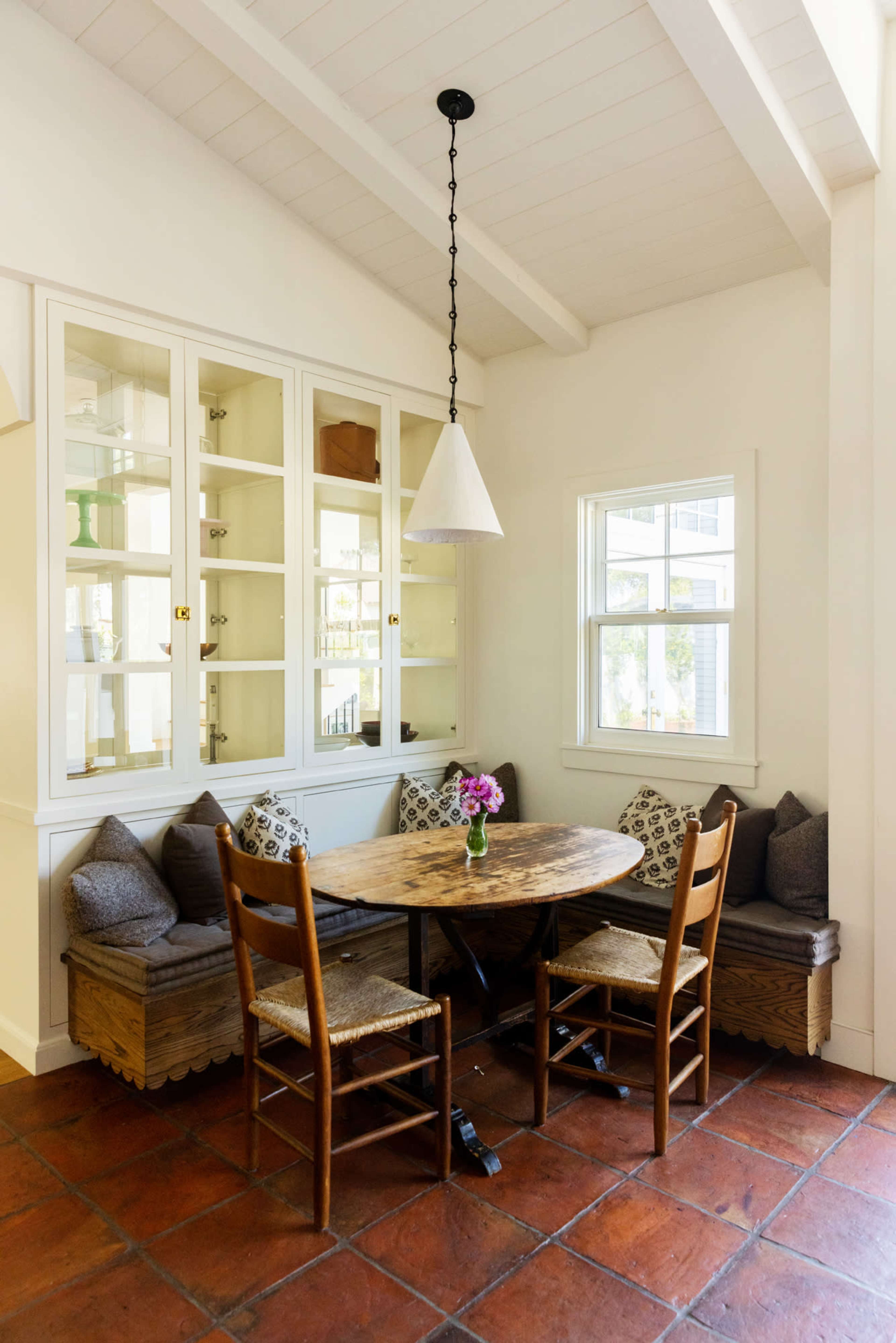 A dining nook features a round table surrounded by two wooden chairs and a built-in bench with pillows, set against a wall of glass-front cabinets.