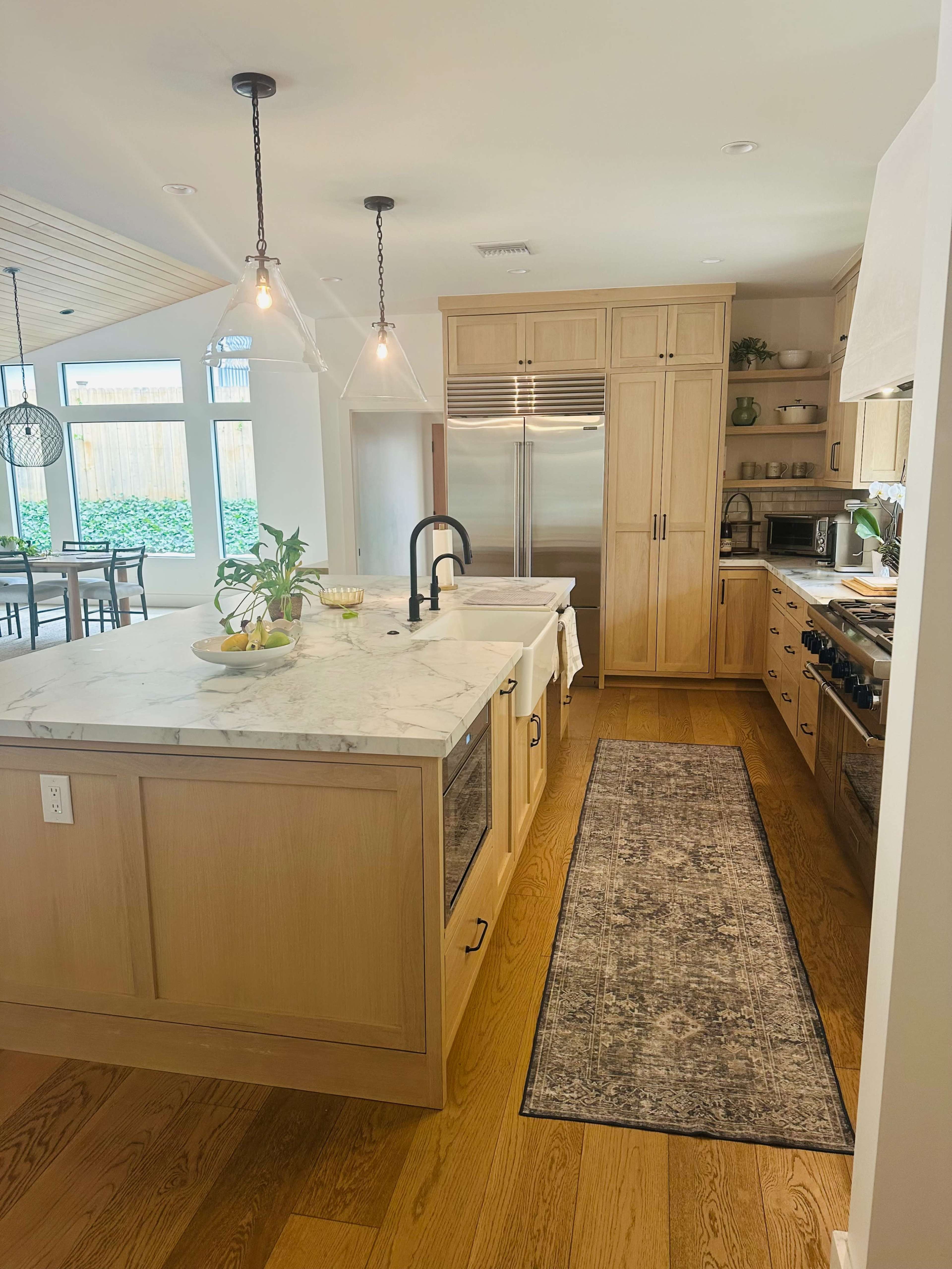 The image shows a modern kitchen featuring a large island with a marble countertop, wooden cabinetry, and stainless steel appliances, illuminated by pendant lights.