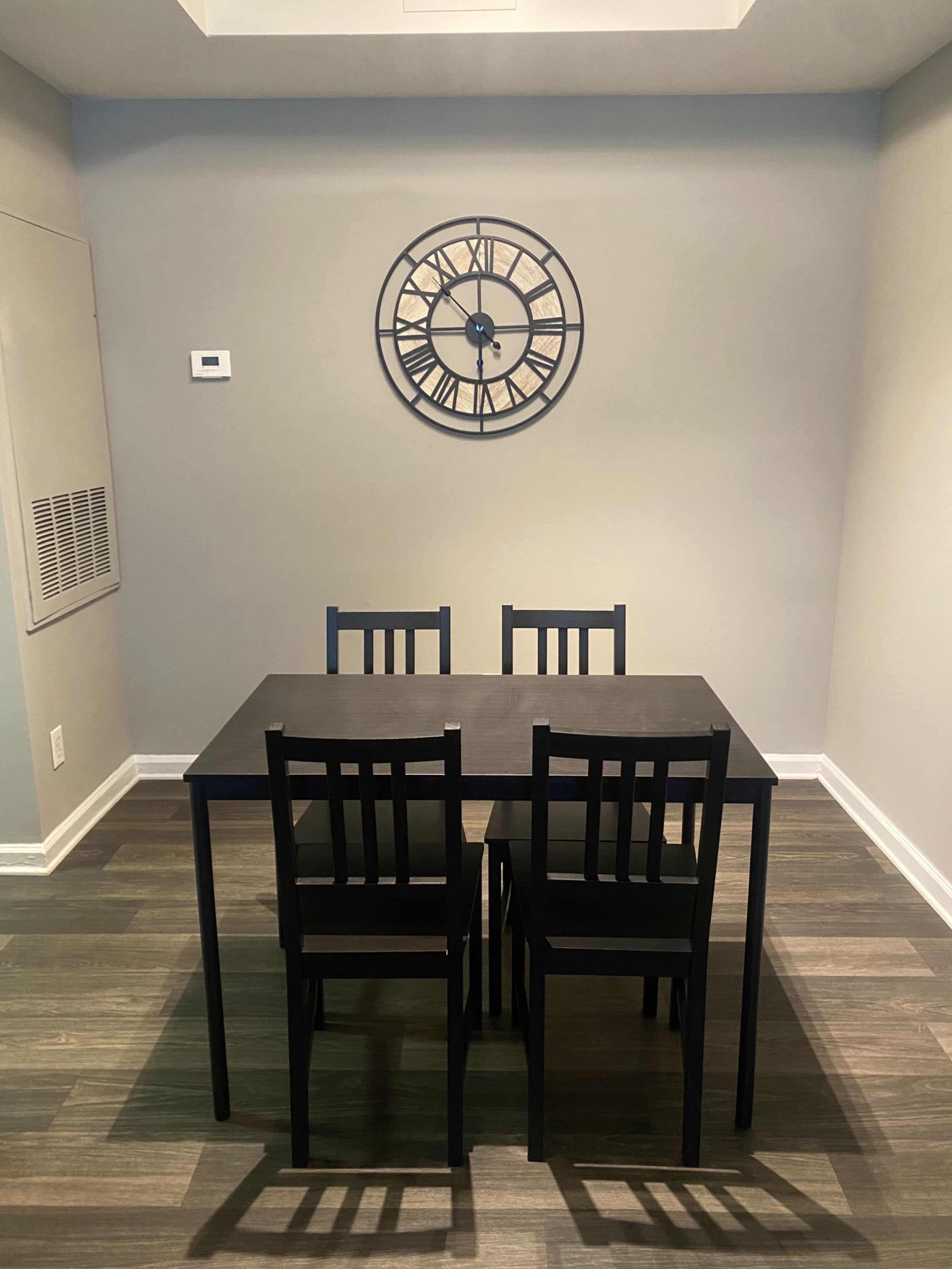 A black dining table with four chairs is positioned in a room featuring a clock on the wall and wooden flooring.