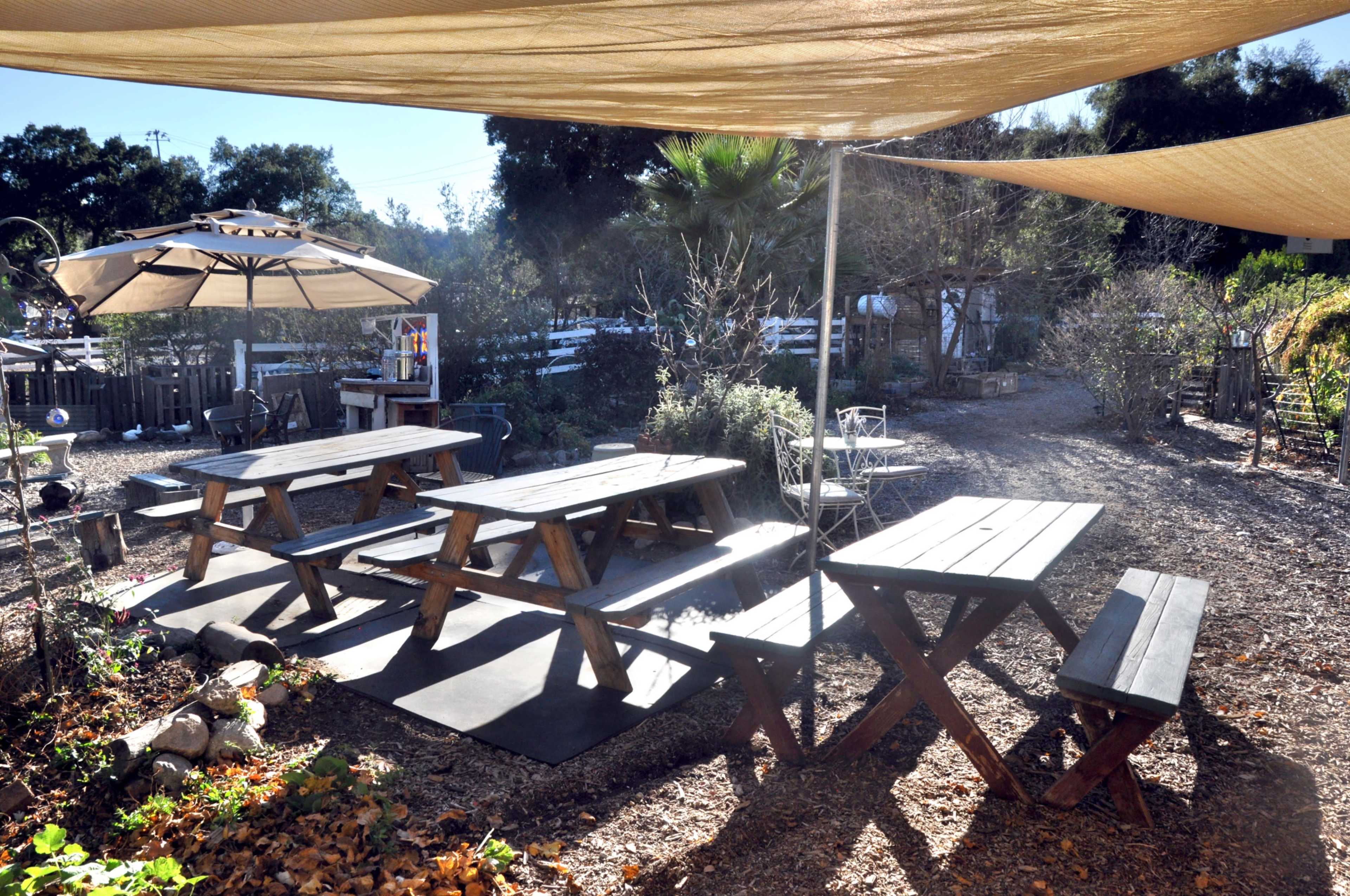 The image shows an outdoor seating area with wooden picnic tables shaded by fabric canopies in a garden setting.