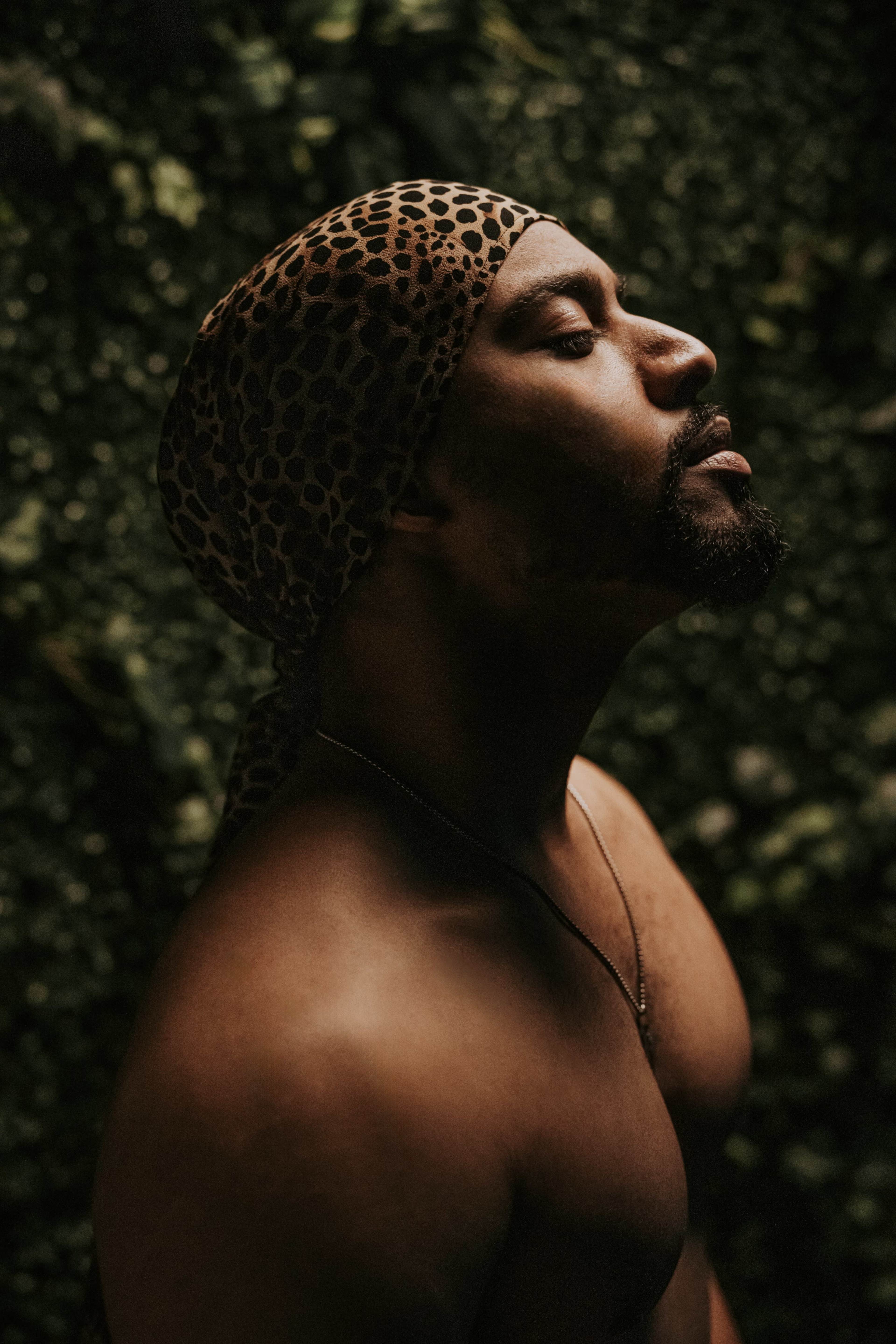A man with a leopard print durag poses against a backdrop of green foliage.