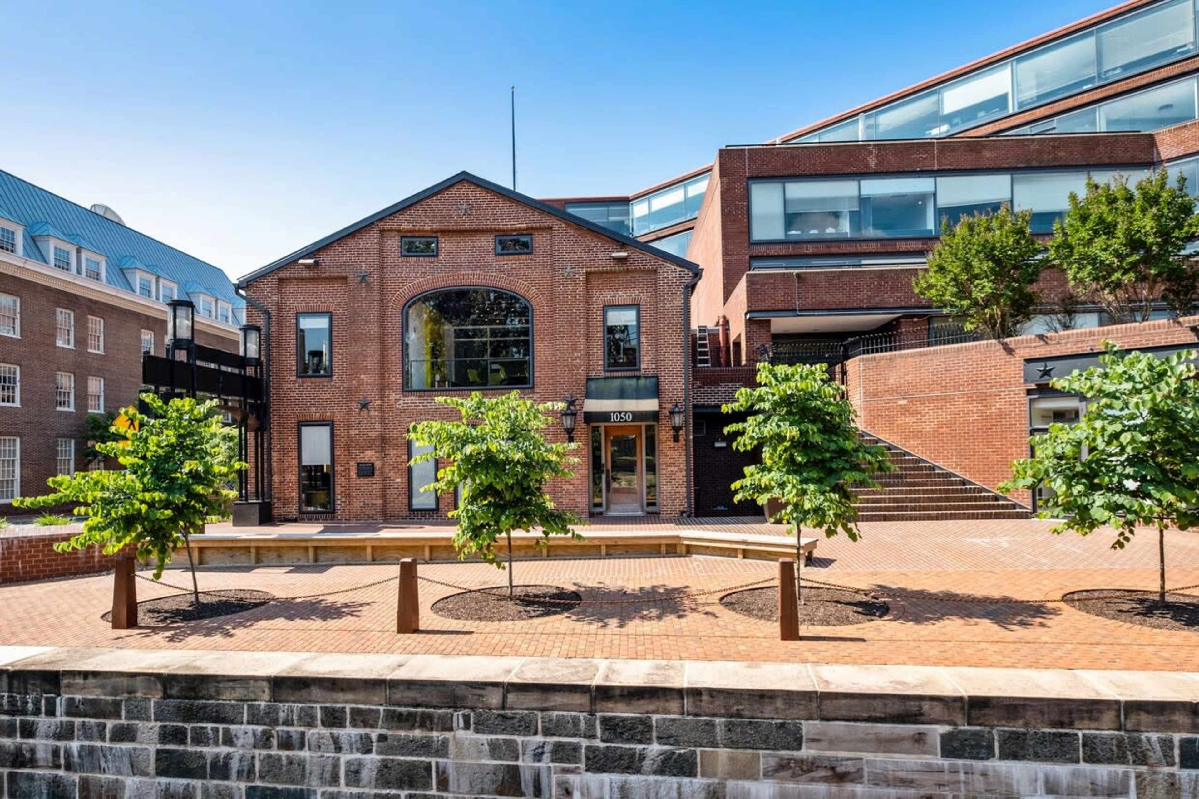 A brick building with large windows set among landscaped walkways and trees, in an urban setting.