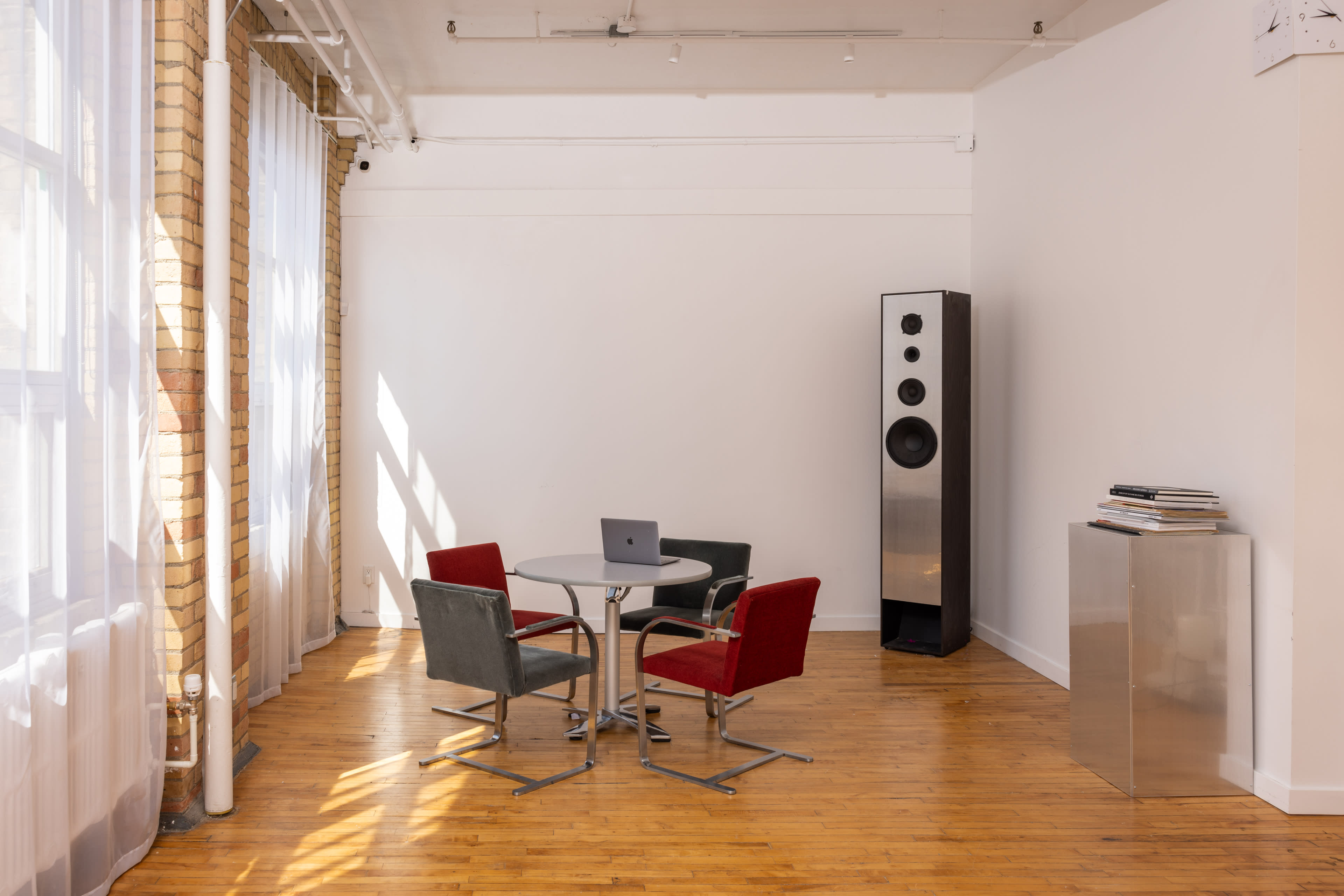 A modern, minimalist interior features a round table with four chairs and a tall speaker against a white wall with large windows letting in natural light.