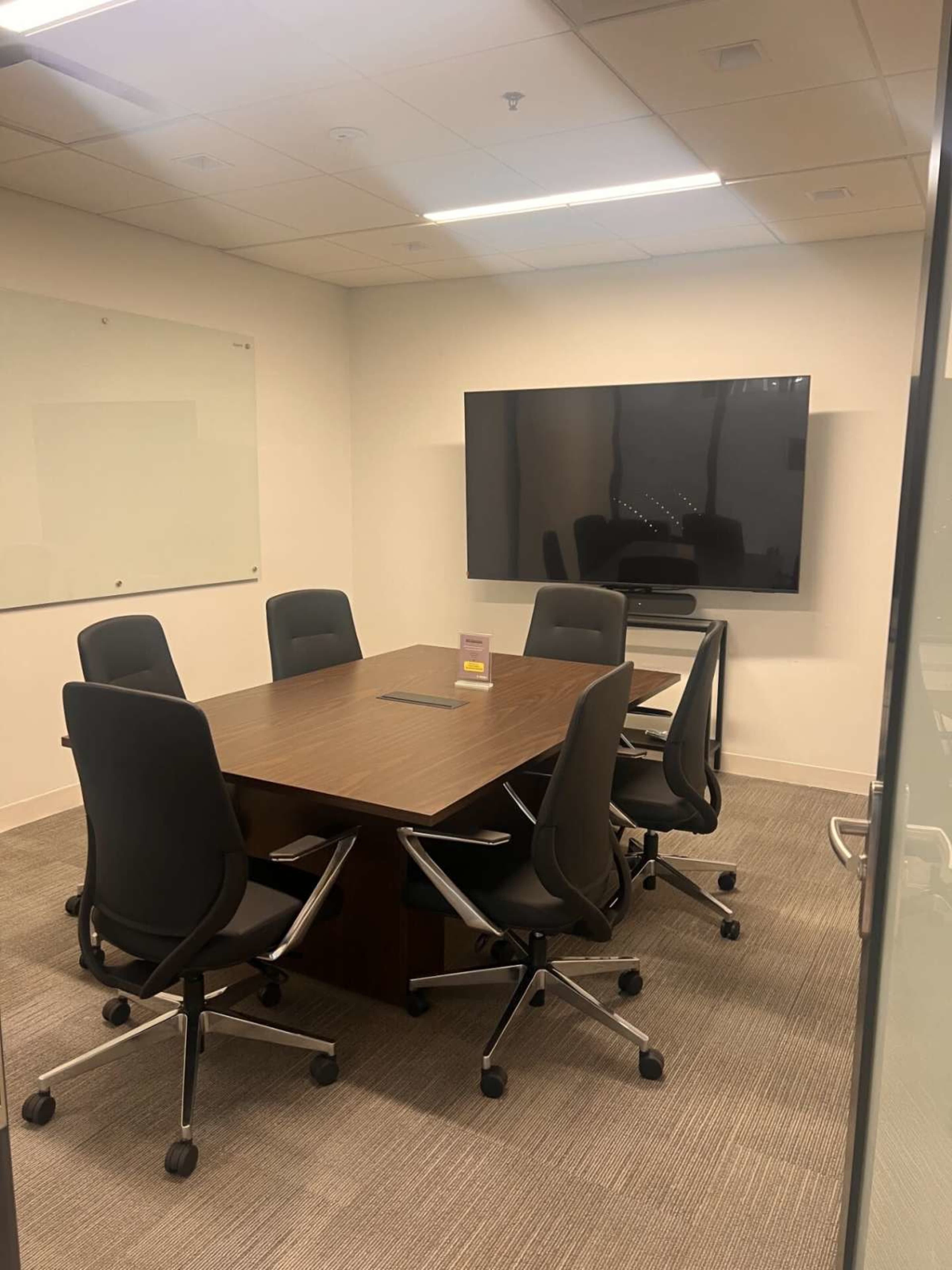 A conference room features a large wooden table surrounded by six black chairs and a wall-mounted television.
