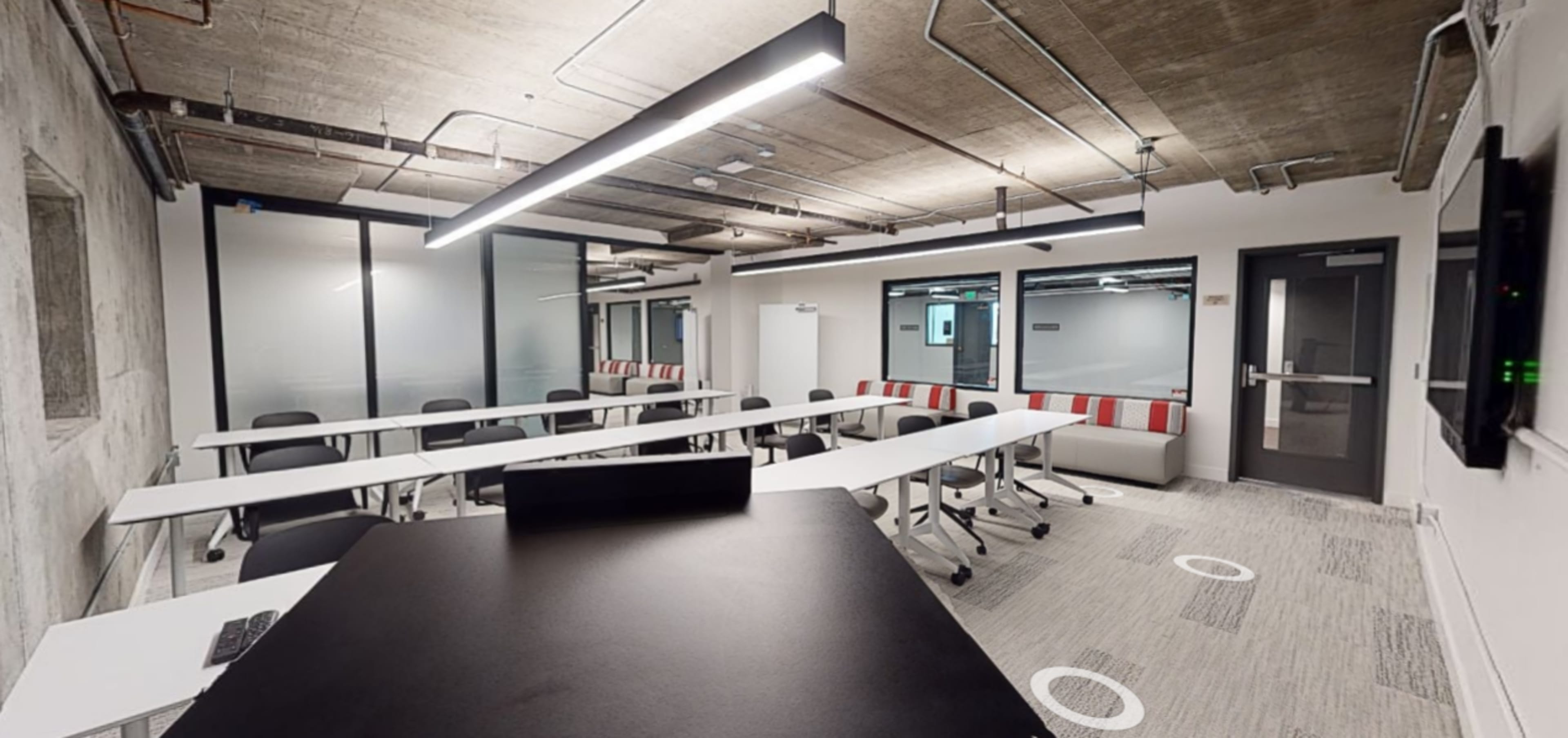 A modern conference room with long tables, black chairs, and a large screen on one wall, featuring a minimalist design and fluorescent lighting.