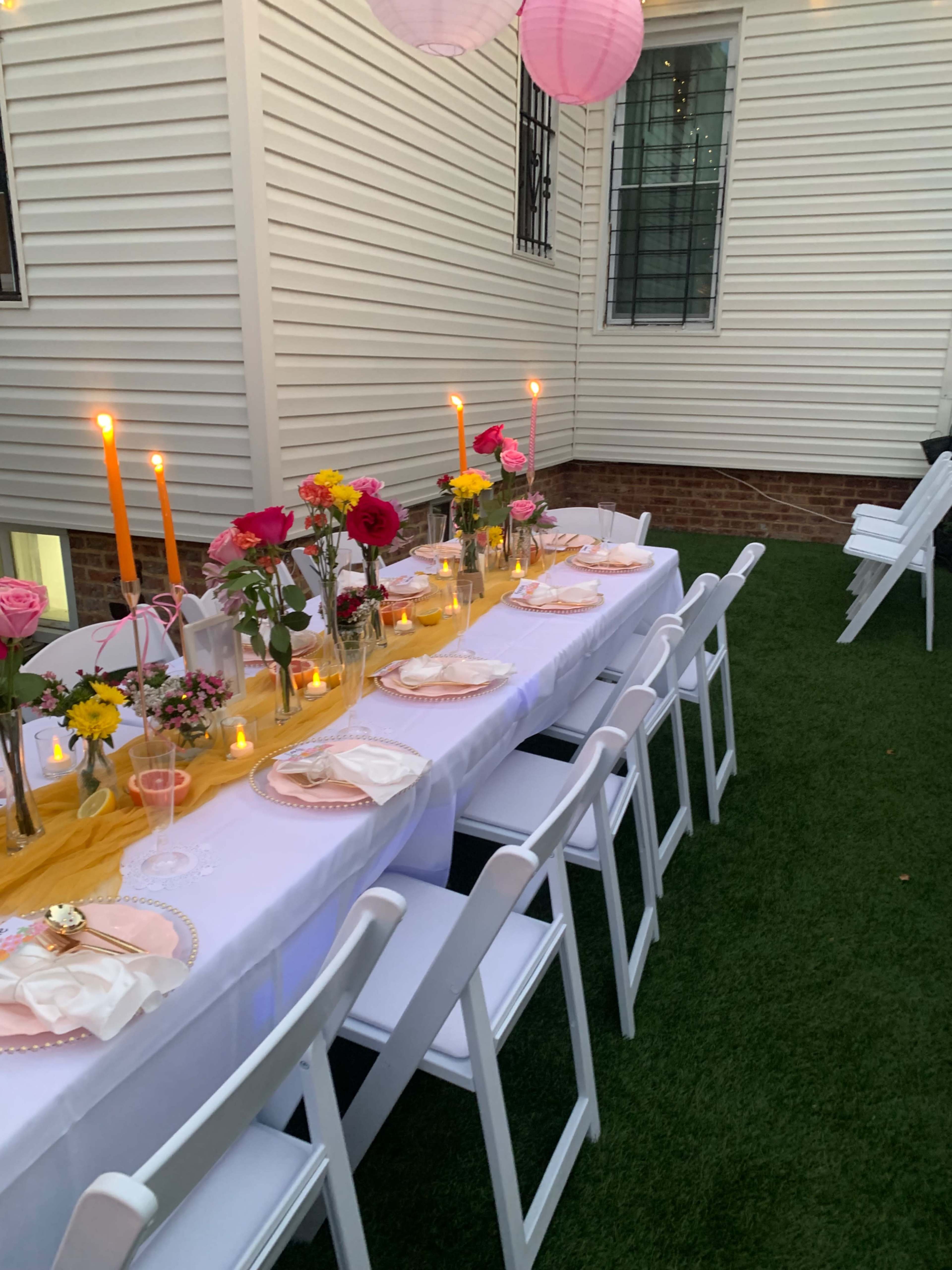 A long table is set outdoors with flowers, candles, and place settings, surrounded by white chairs on a green lawn.