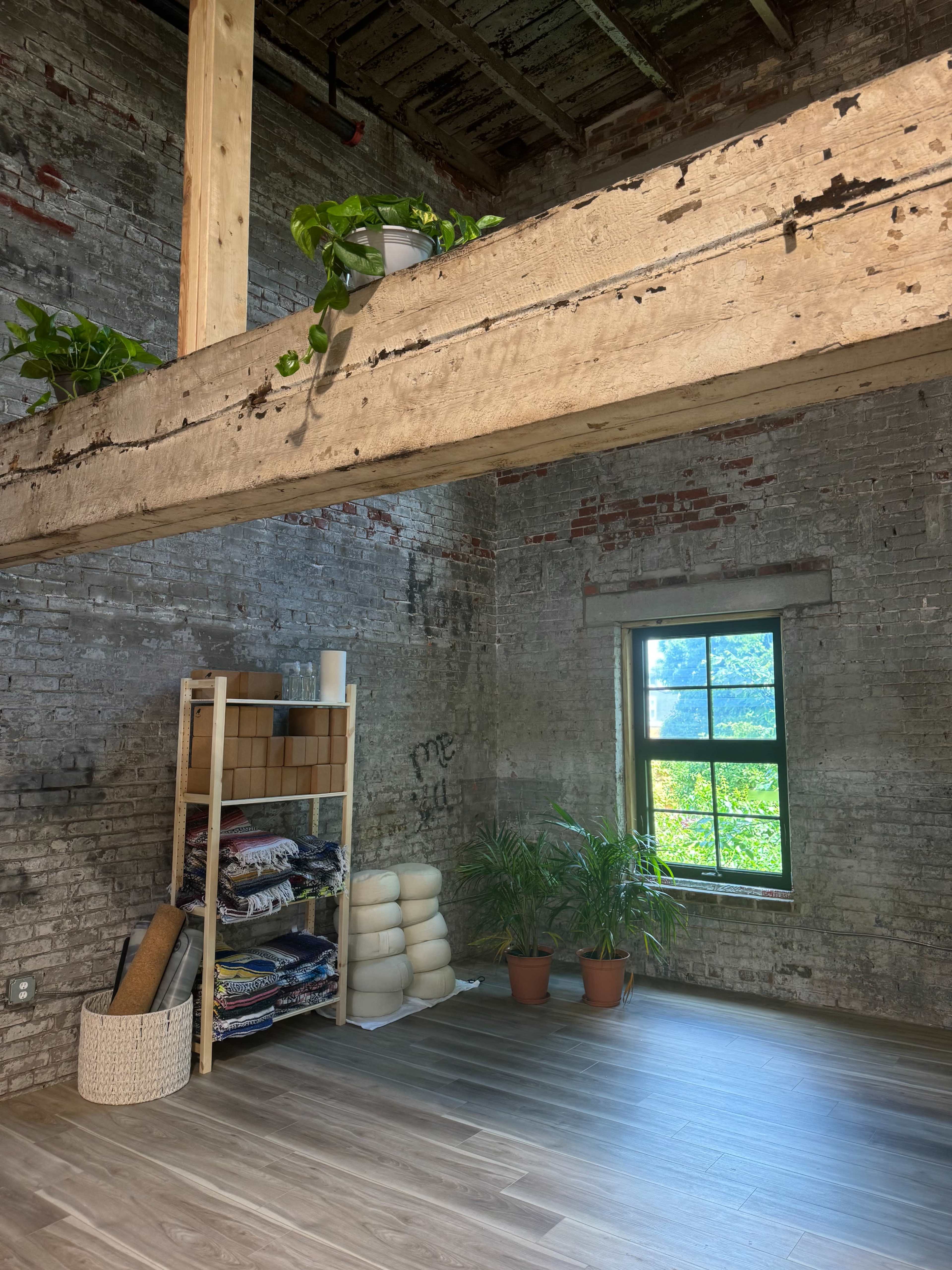 An industrial-style room with exposed brick walls, a wooden beam above, a shelving unit with rolled textiles, and potted plants beside a window.
