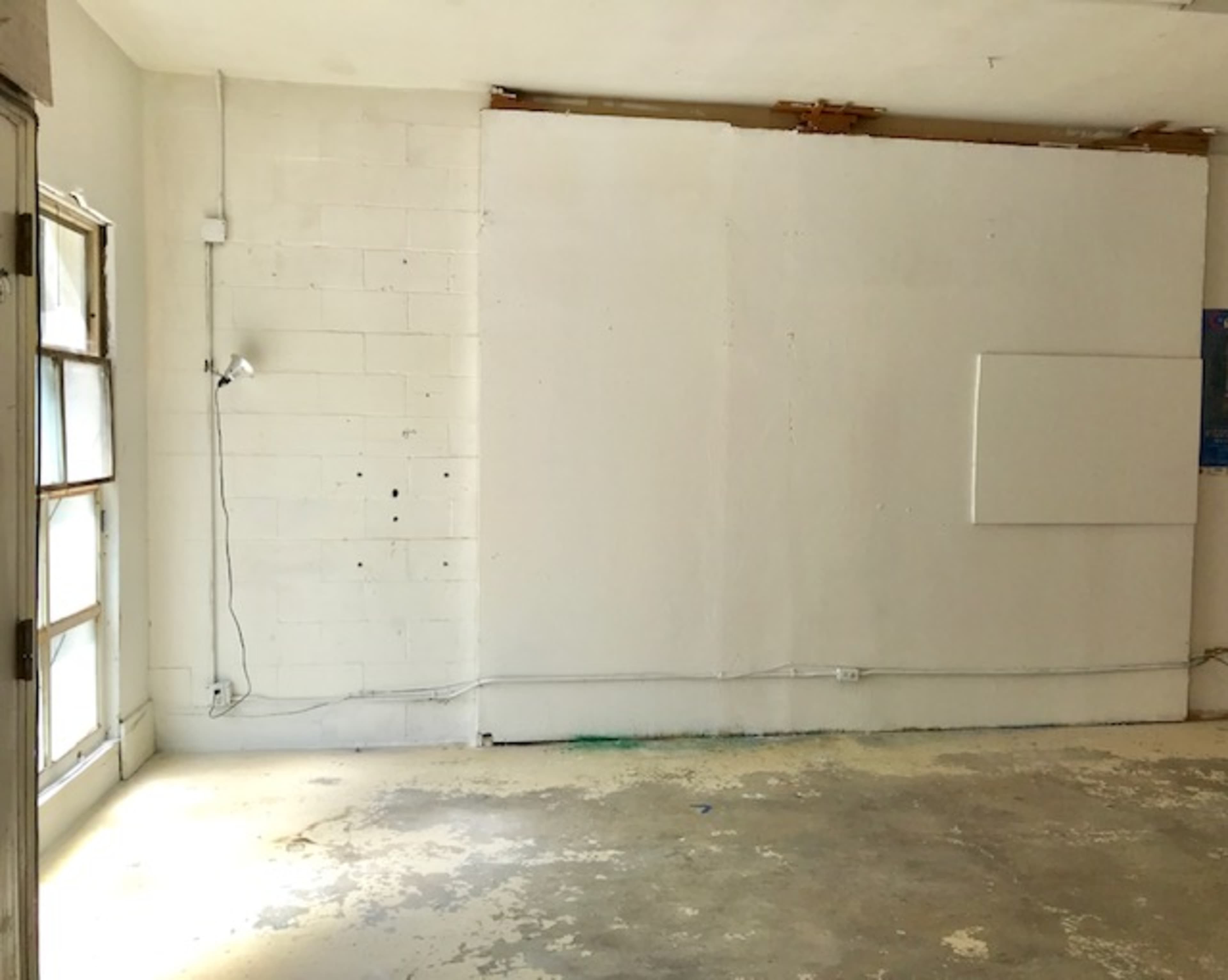 An empty room features a bare white wall with visible marks and a small light fixture attached, along with patchy flooring.