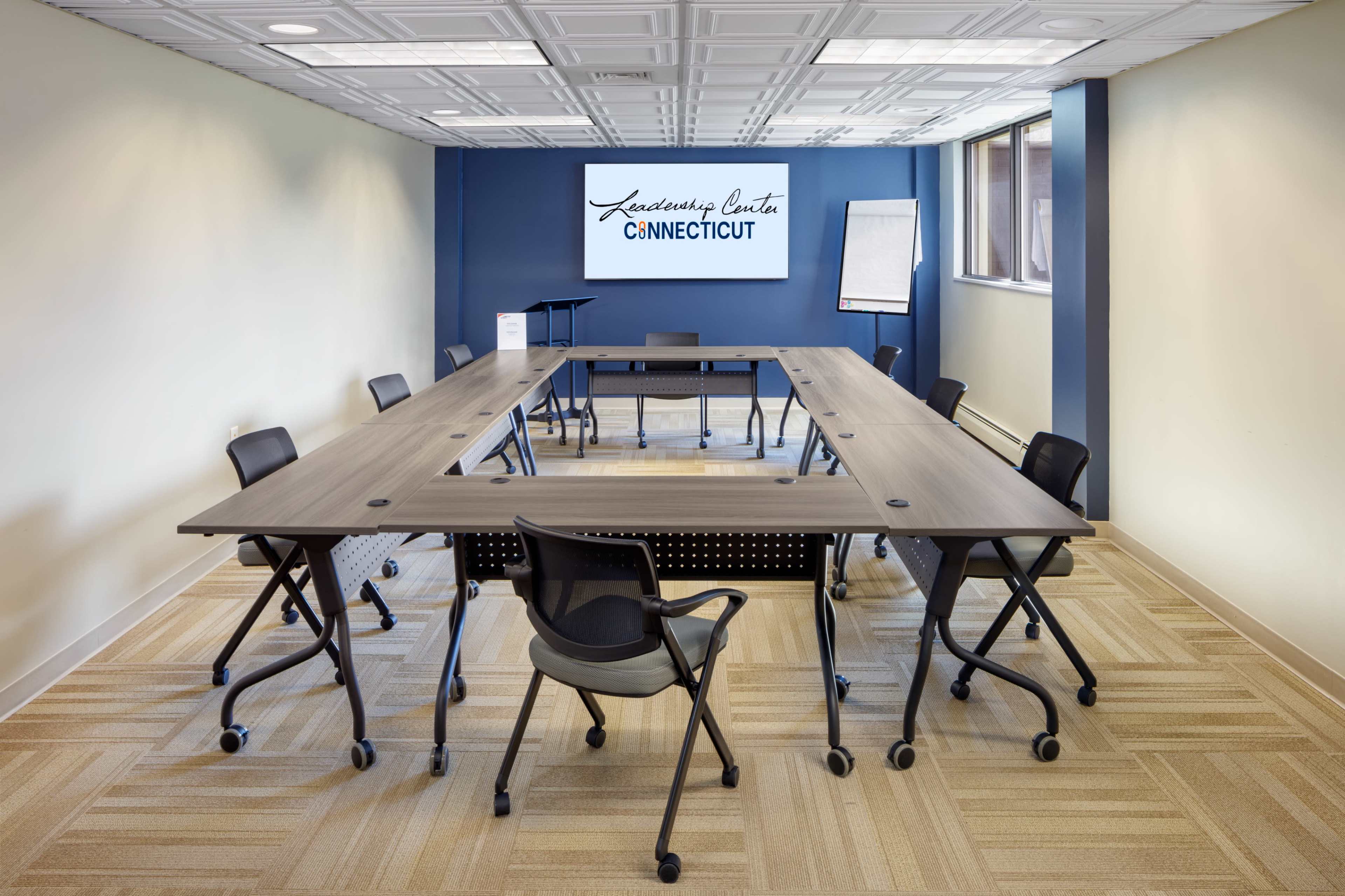 A modern conference room features a large, rectangular table surrounded by rolling chairs, with a presentation area and a sign that reads "Leadership Center Connecticut" on the wall.