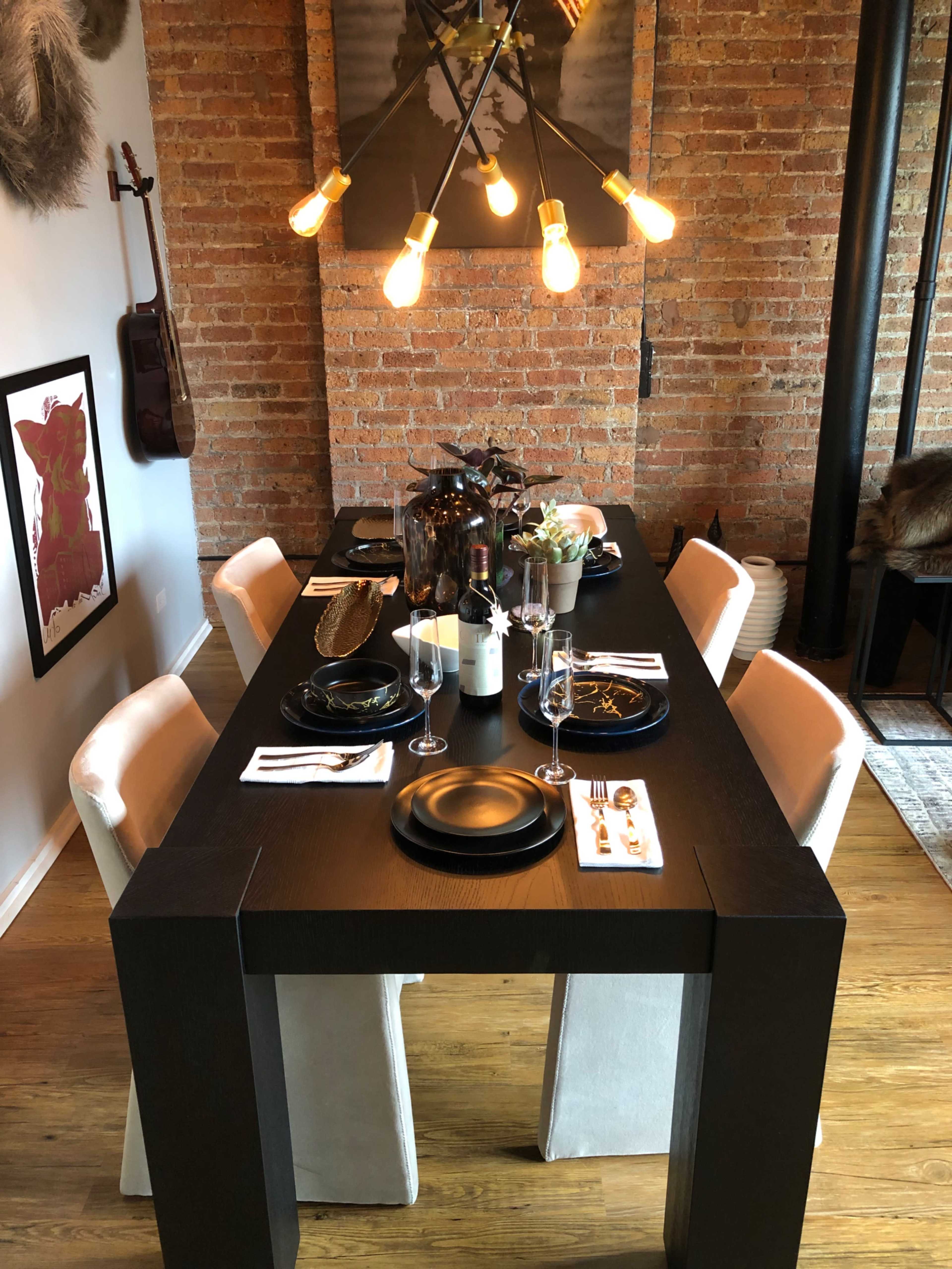 A long, rectangular dining table is set for a meal with black plates, wine glasses, and decorative centerpieces, under a stylish light fixture in a room with exposed brick walls.
