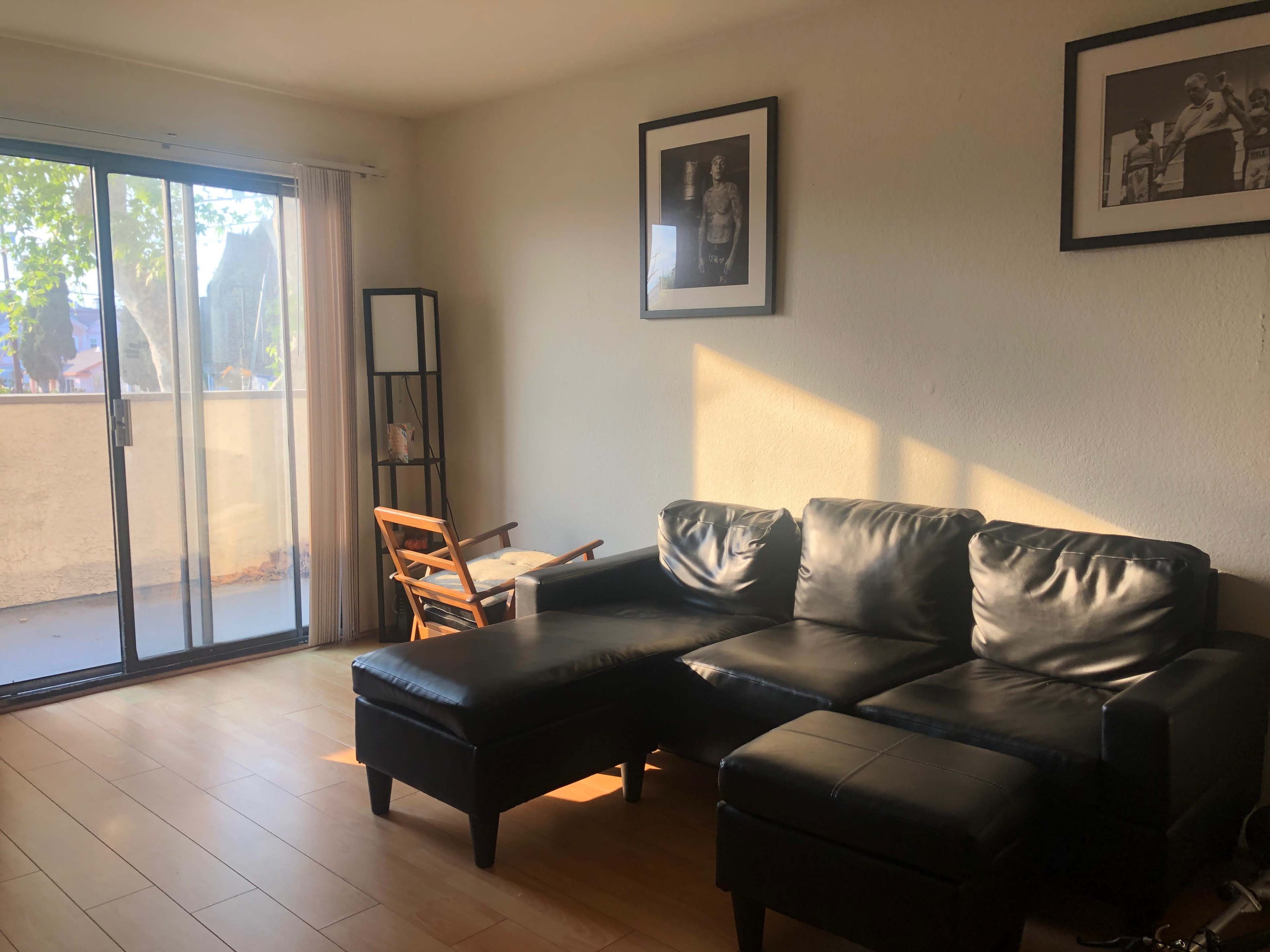 The living room features a black leather sectional sofa, a small wooden chair, and large windows illuminating the space with natural light.