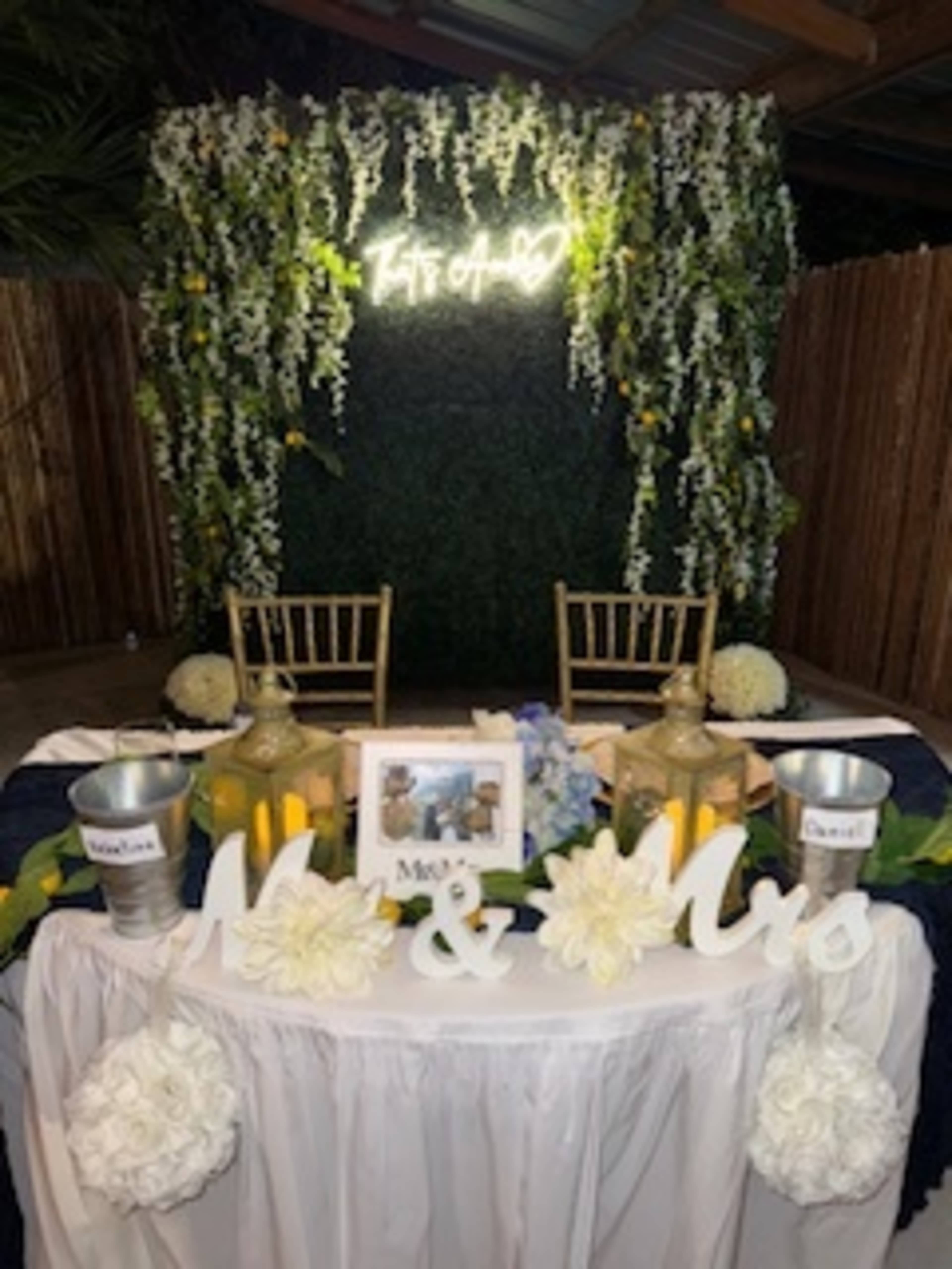 The image shows a decorated reception area with a table for the bride and groom, featuring floral arrangements, a lighted sign, and two gold lanterns.