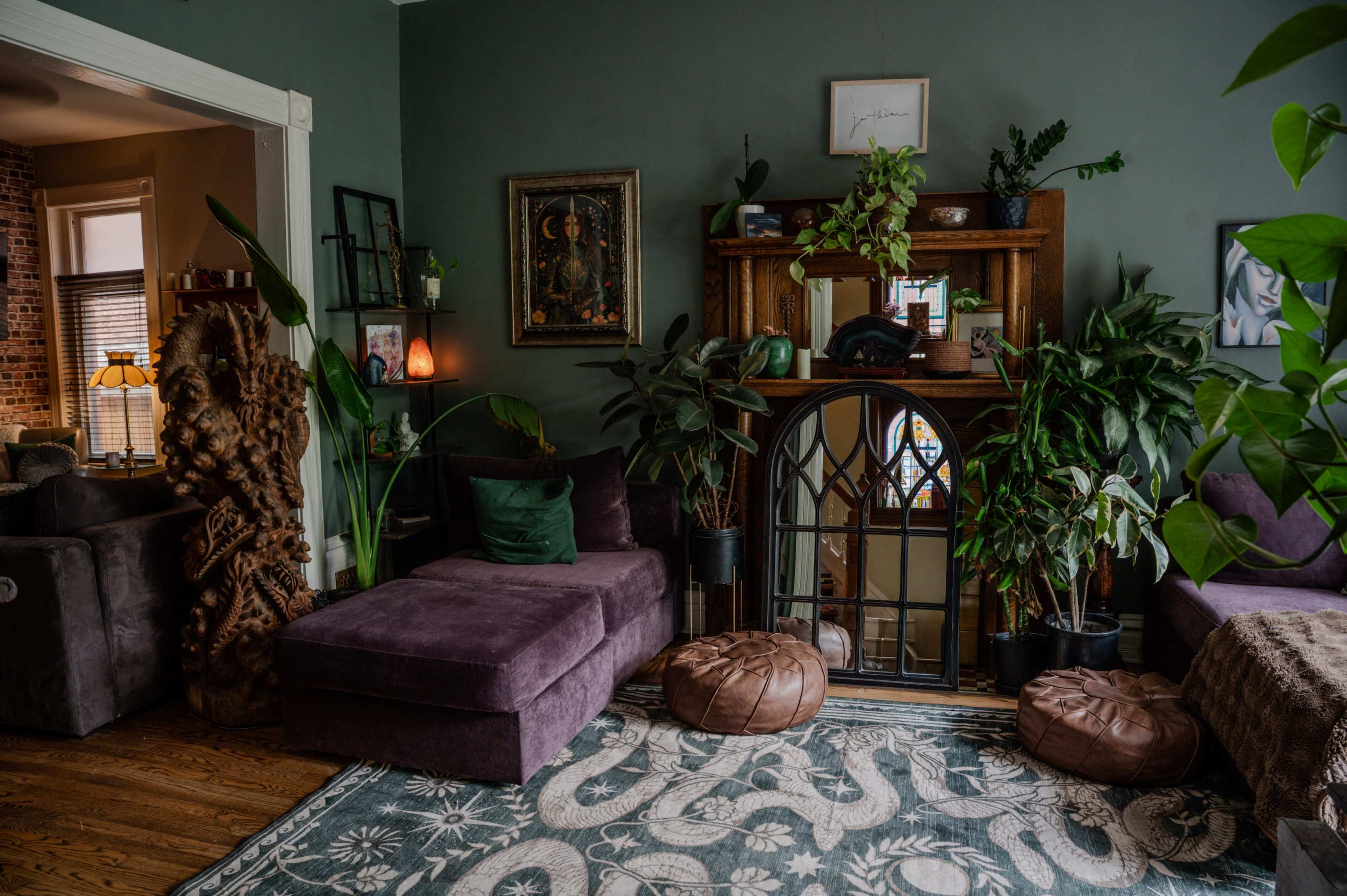 The image shows a cozy living room featuring a purple sectional couch, greenery, and a decorative mirror above a wooden mantle.
