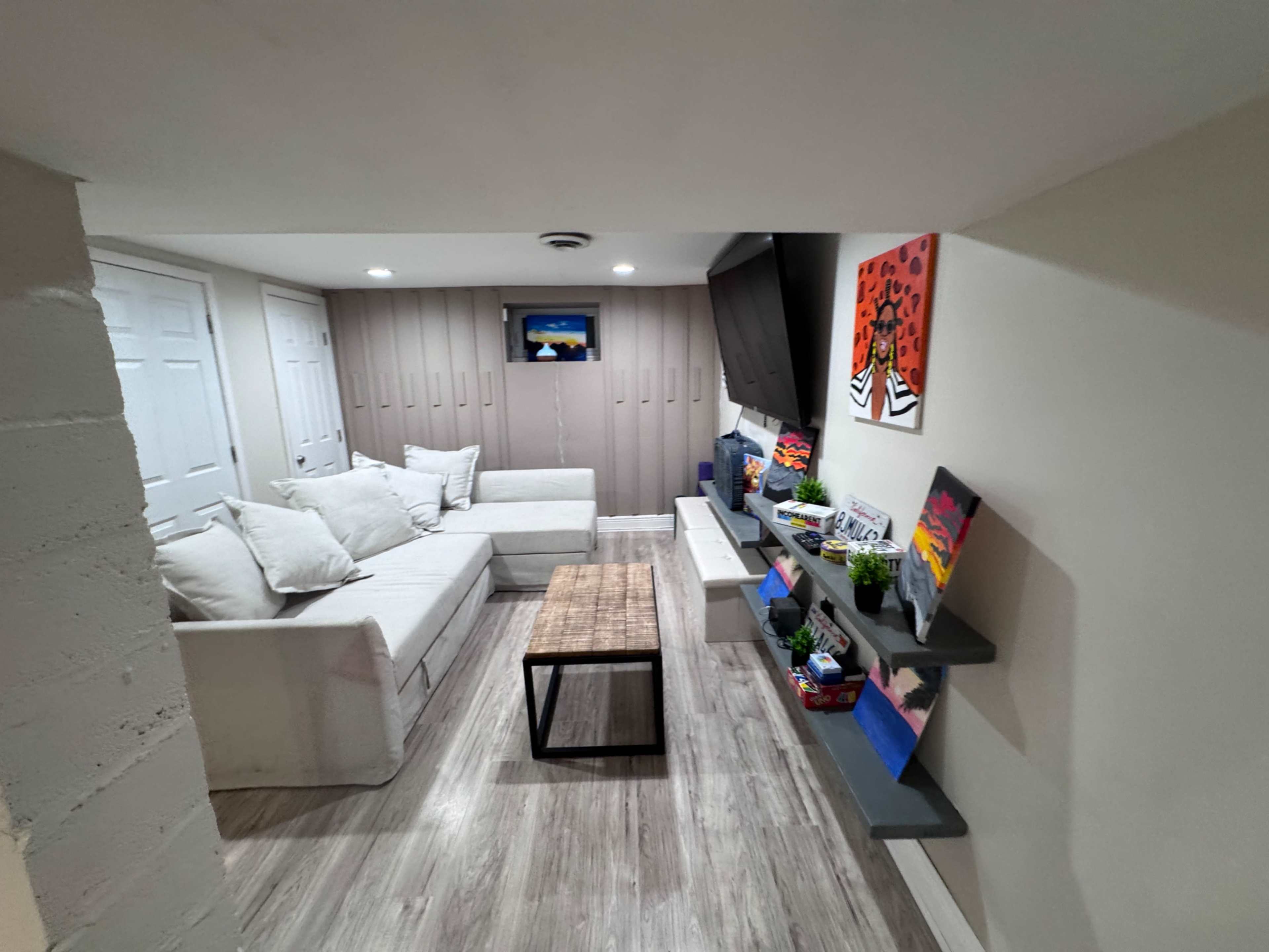 The image shows a basement living area with a sectional couch, a wooden coffee table, and wall-mounted shelves displaying decorative items and plants.