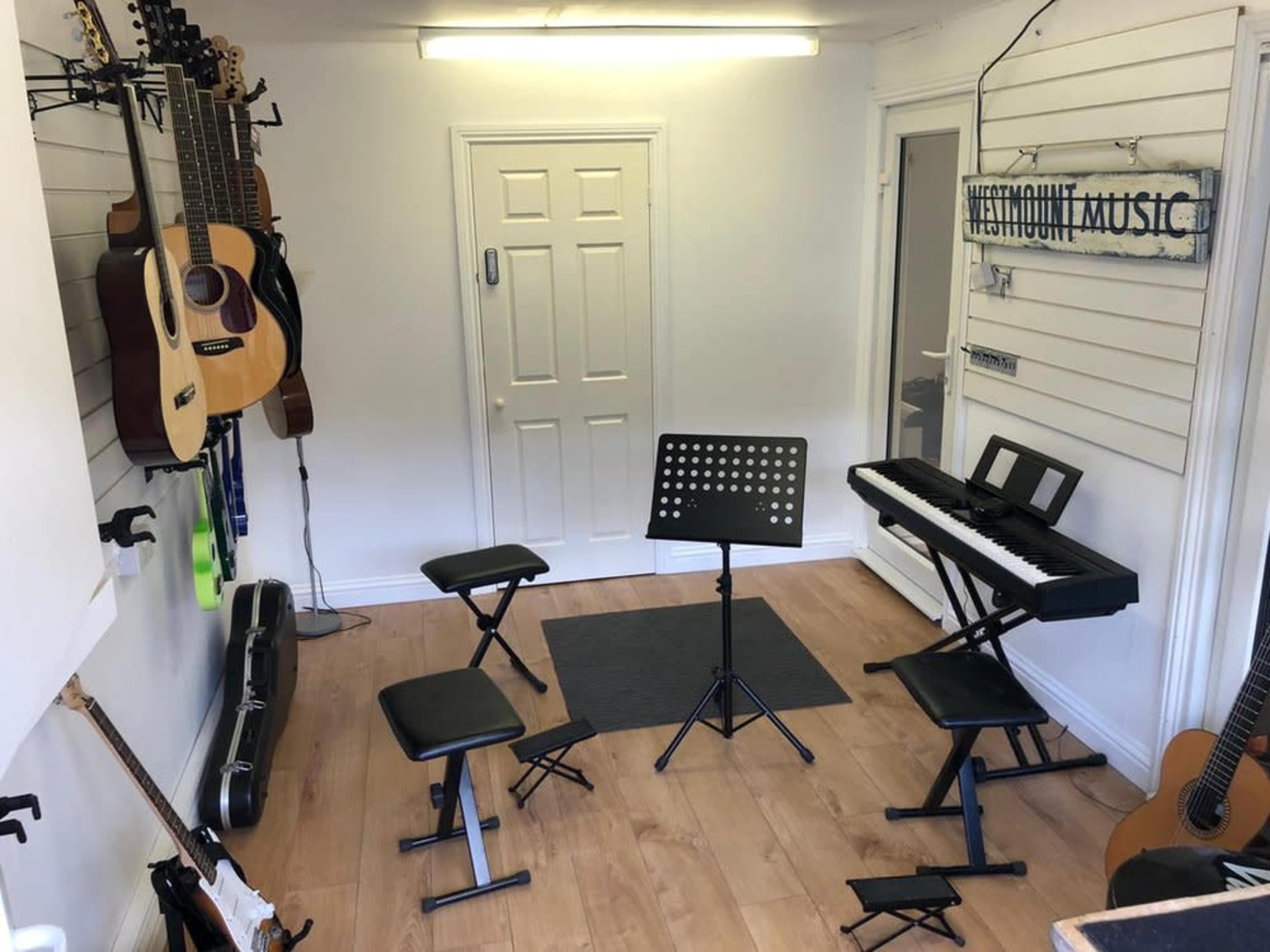 The image shows a small music room with several guitars on the walls, a keyboard on a stand, and four music stools arranged around a music stand.