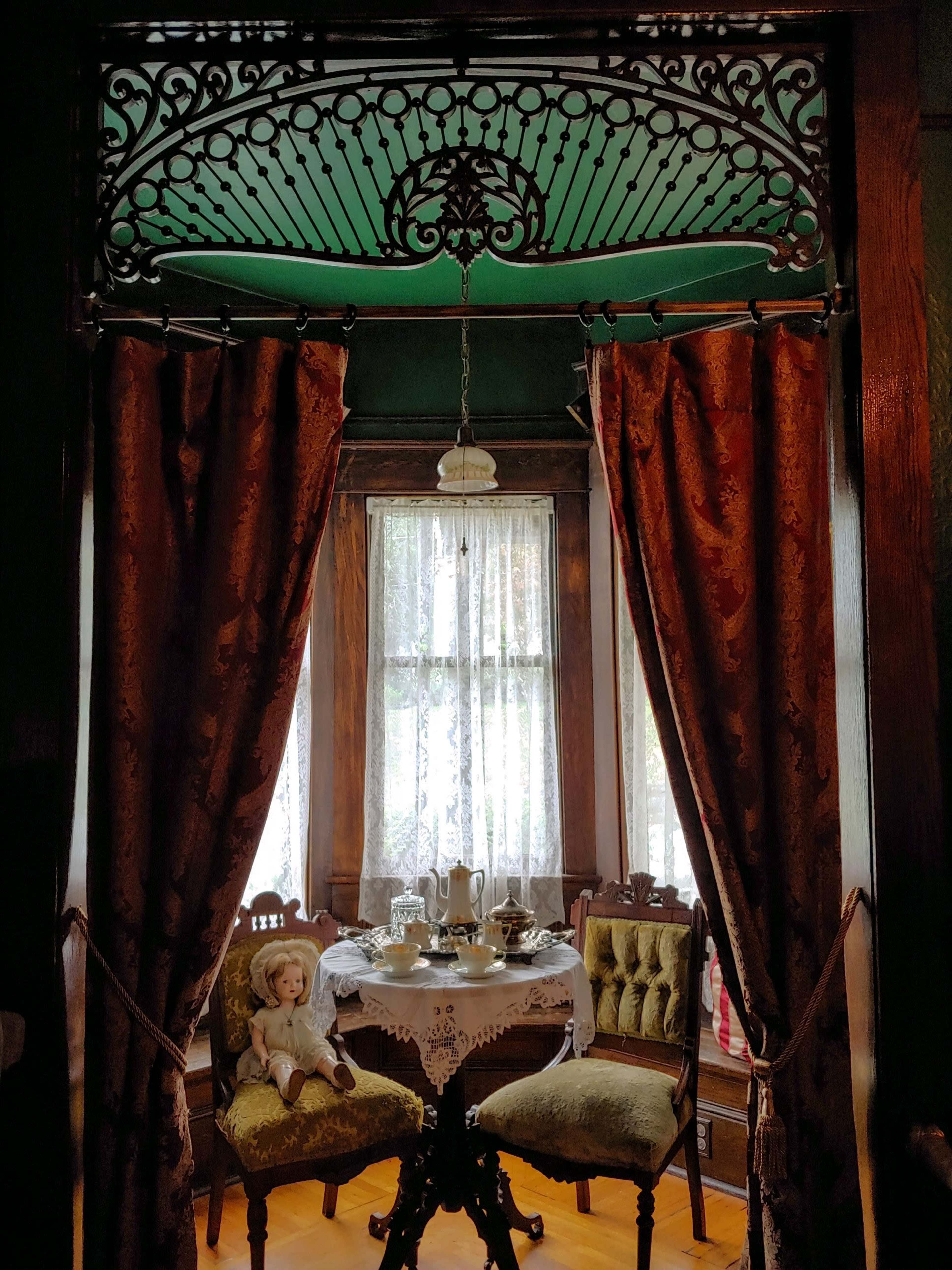 A small vintage dining area is framed by red curtains, featuring a table set for tea and two ornate chairs, with a doll placed on one of the seats.