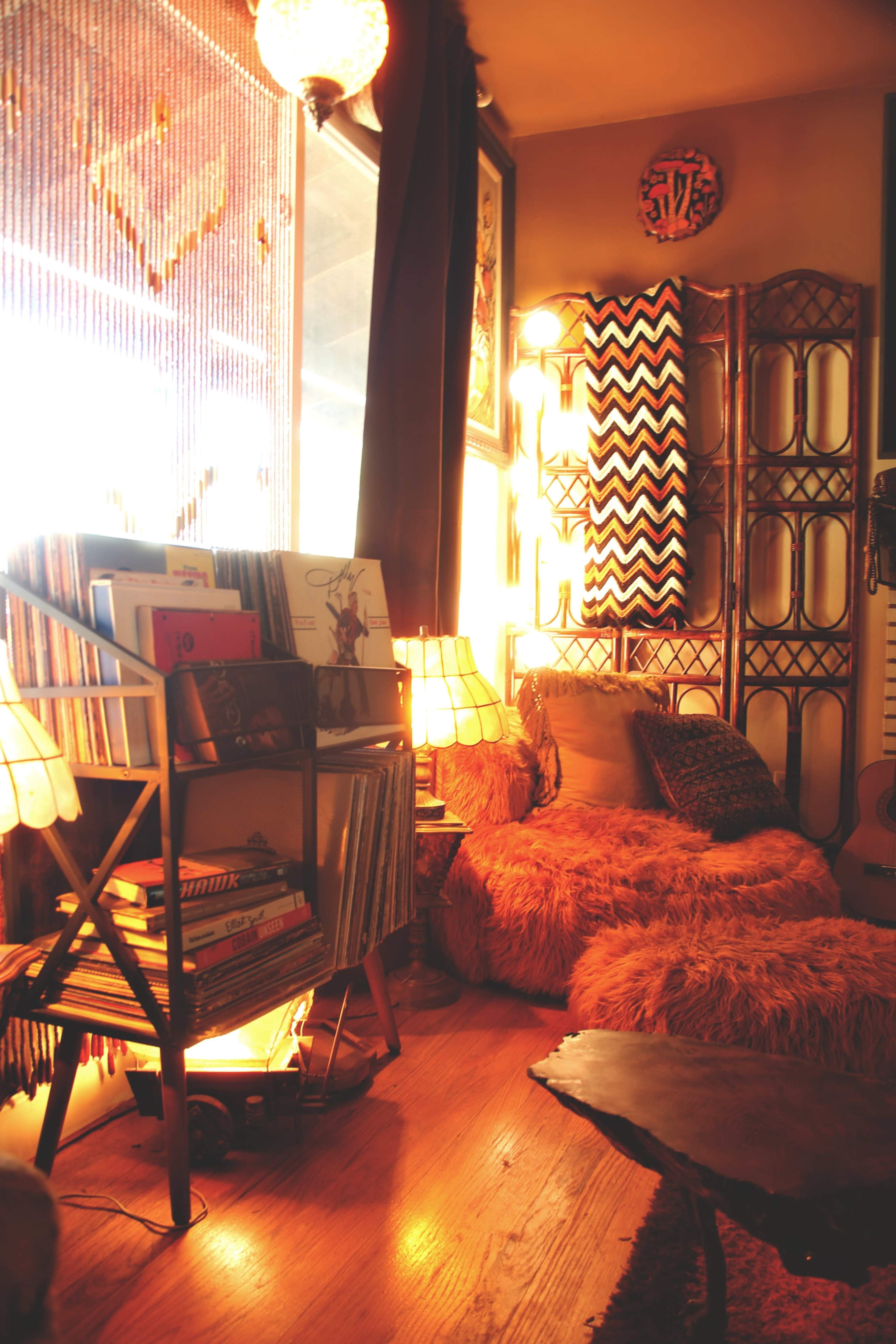 The image shows a cozy corner of a room featuring a shelving unit filled with records, a plush seating area covered in fur-like fabric, and warm lighting from lamps.
