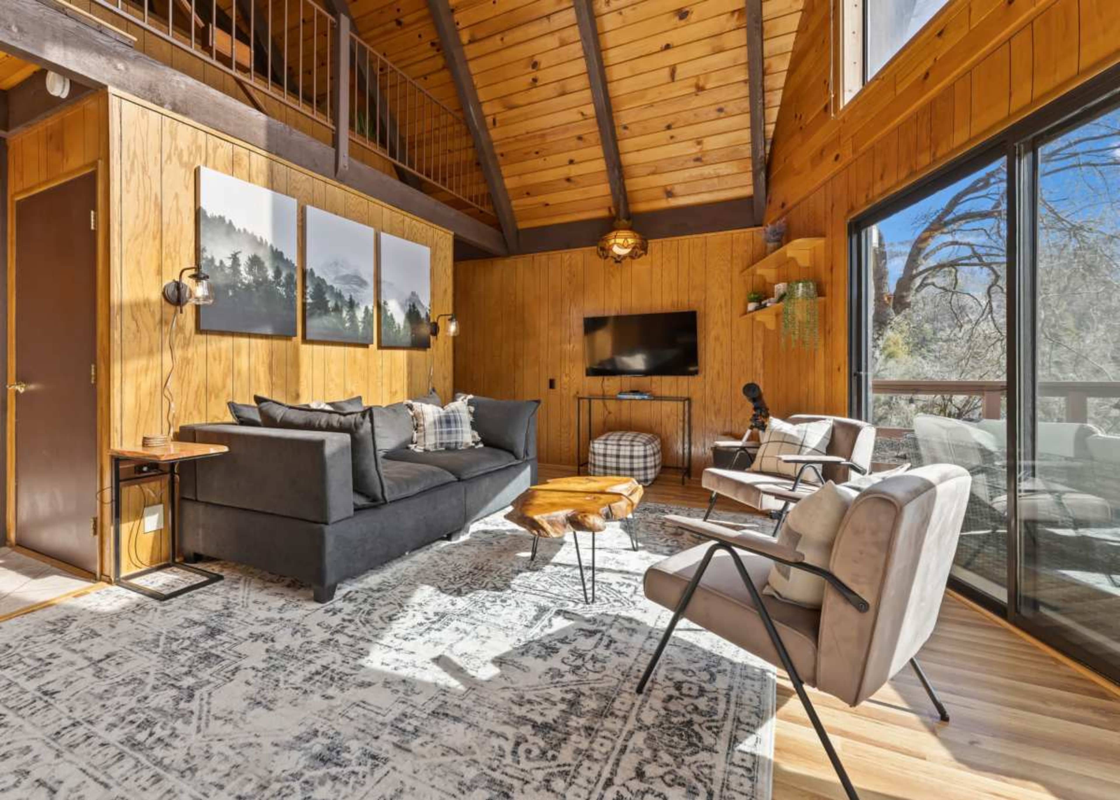 A cozy living room with wooden walls, a high ceiling, a sectional sofa, two chairs, a television, and large windows providing natural light.