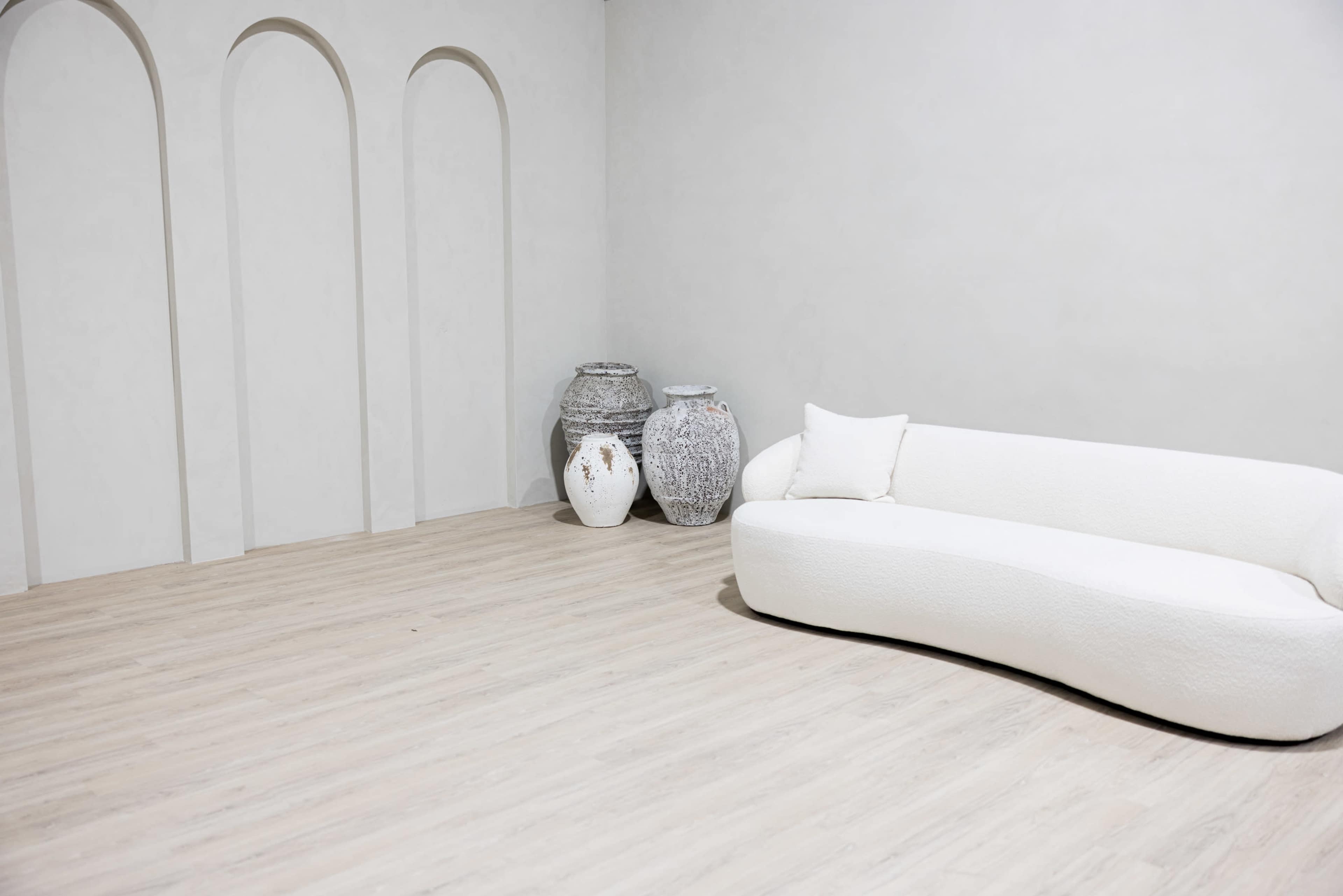 A curved white sofa is positioned next to three decorative vases in a minimalist room with arched wall niches.