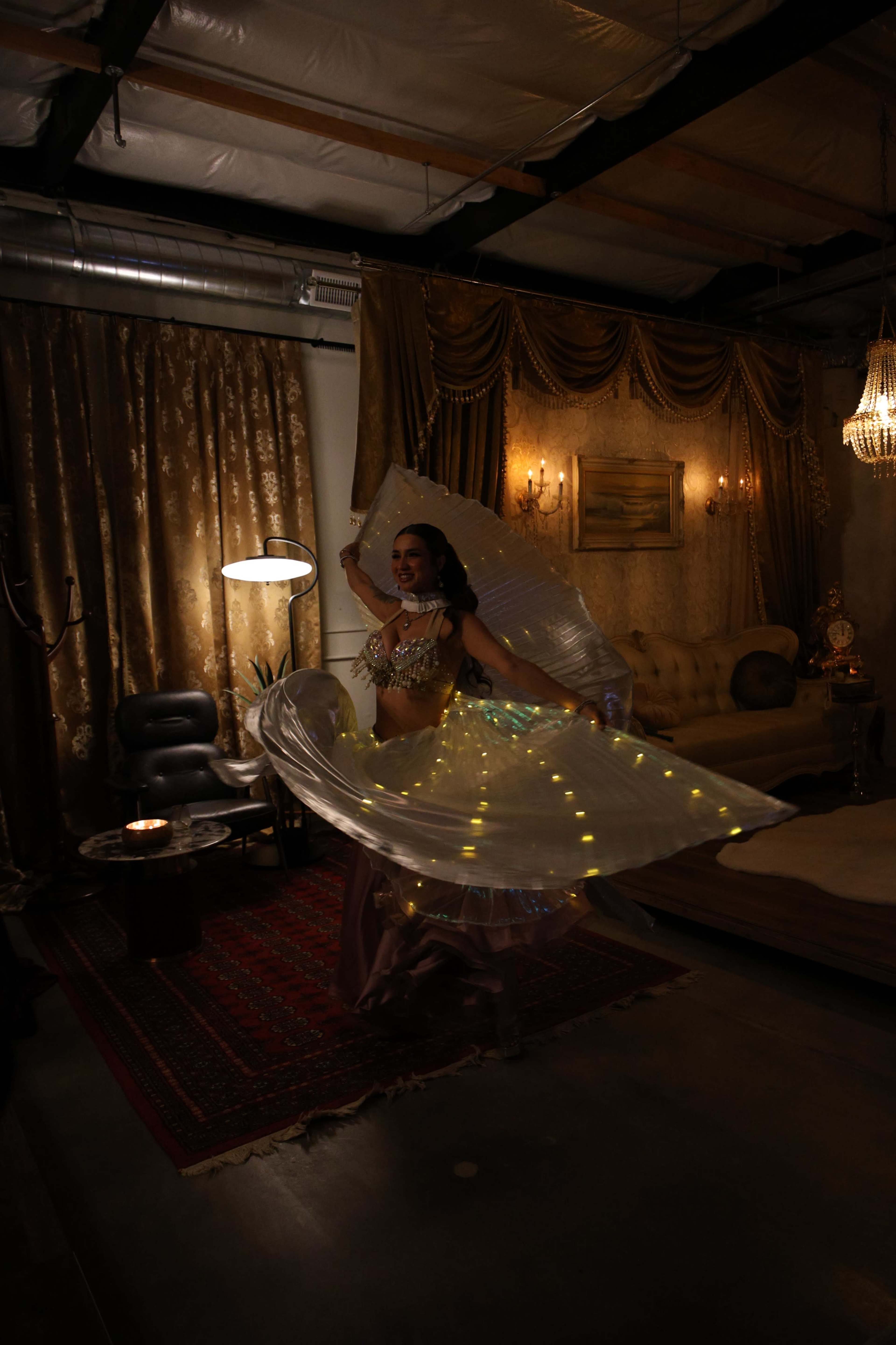 A dancer twirls in a dimly lit room, wearing a sparkling costume and a flowing fabric adorned with lights.