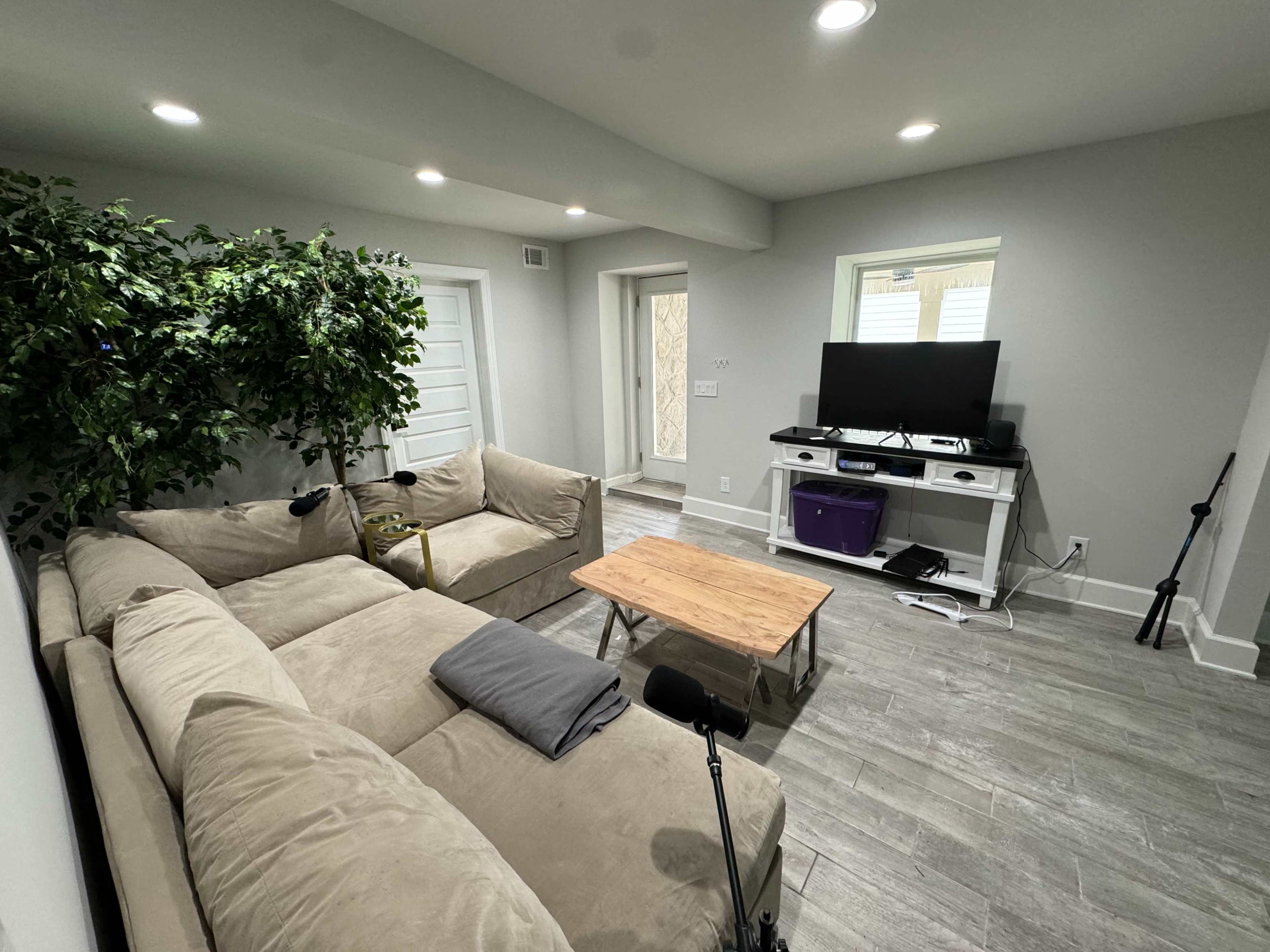 A living room features a sectional sofa, a wooden coffee table, a TV stand with a television, and a potted tree in the corner.