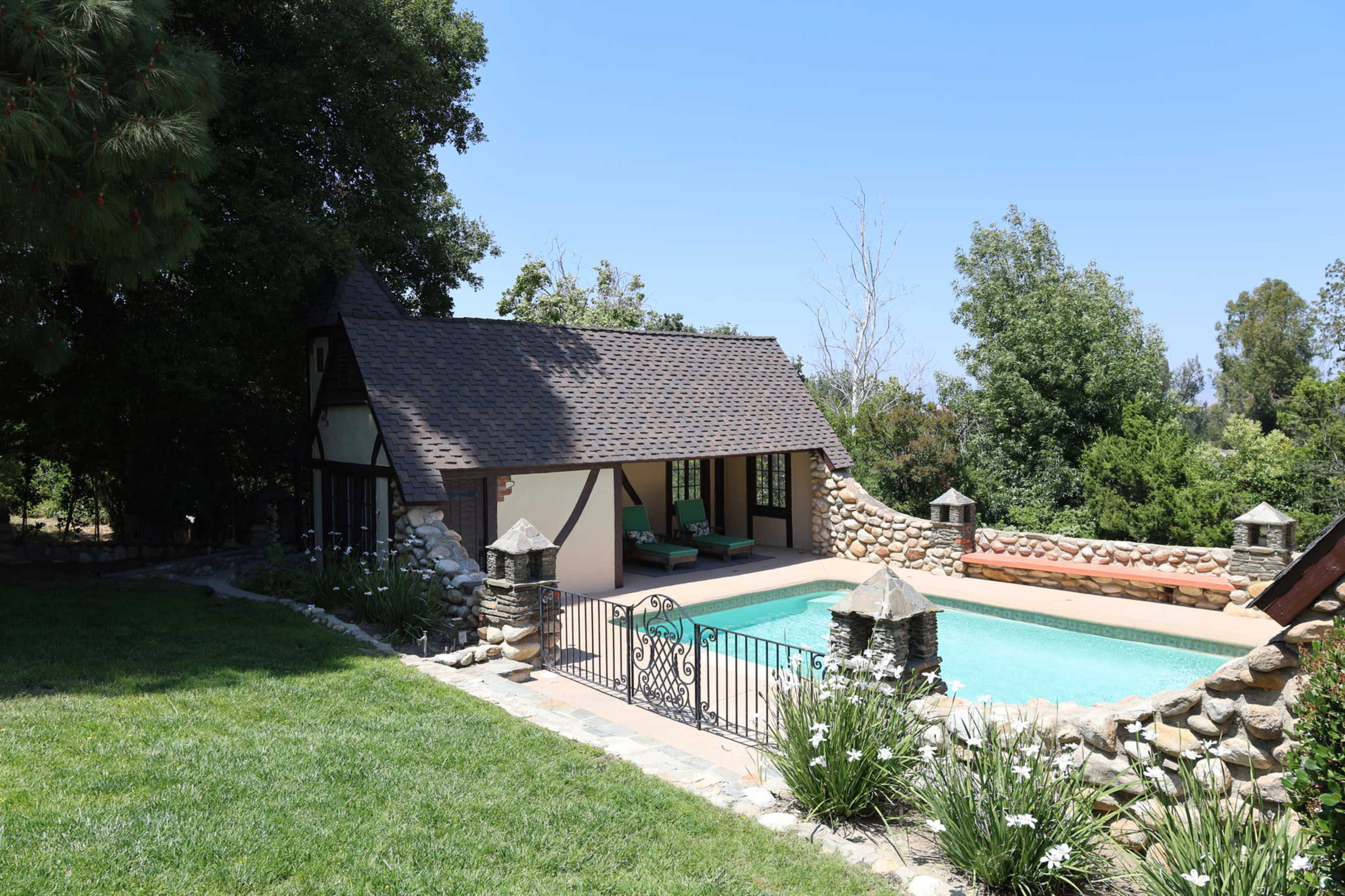 A small, rustic house with a steep roof is situated beside a swimming pool, surrounded by stone walls and lush greenery.