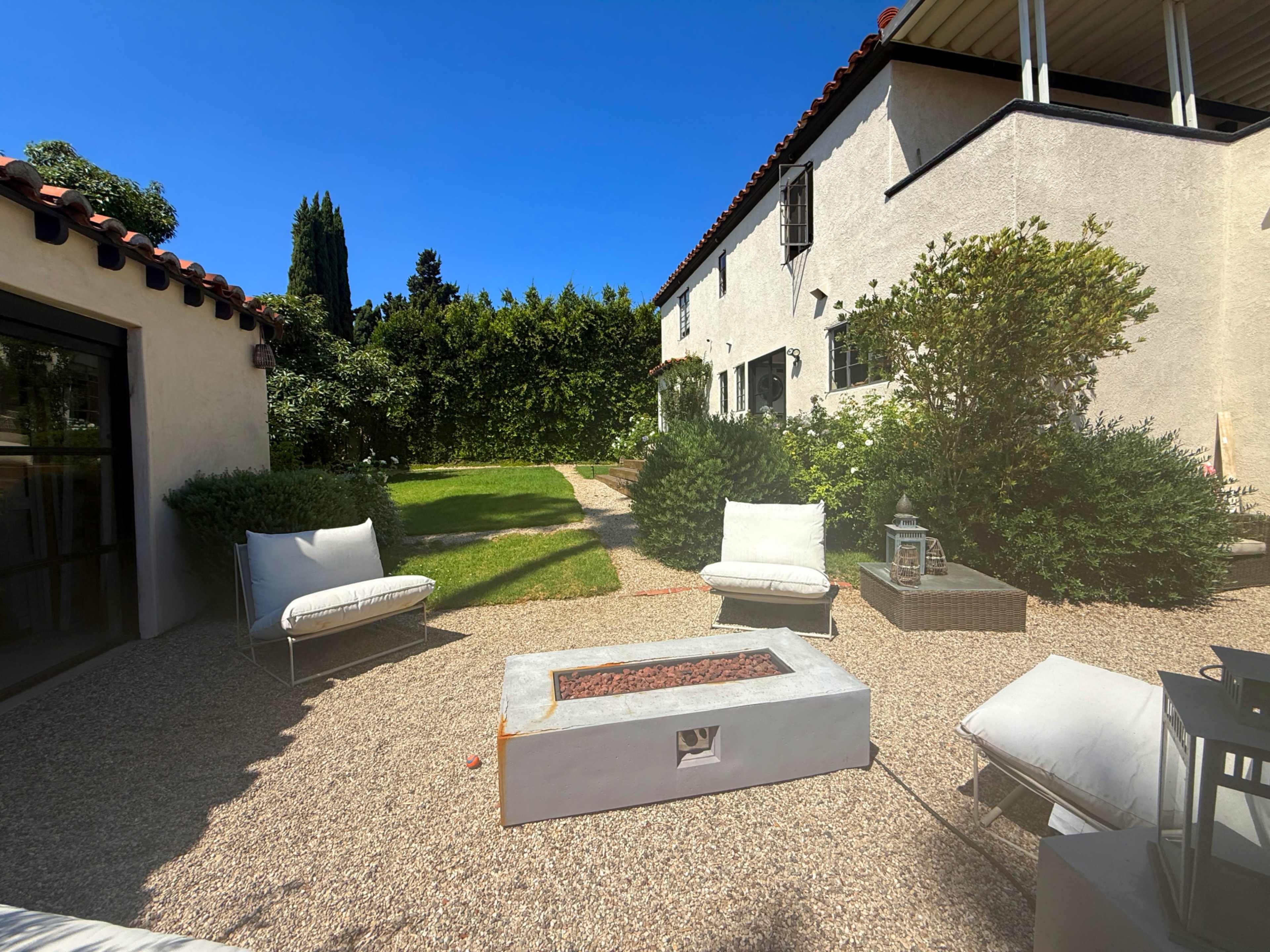A landscaped outdoor area features a gravel surface with two white lounge chairs, a concrete fire pit, and greenery surrounding a lawn.
