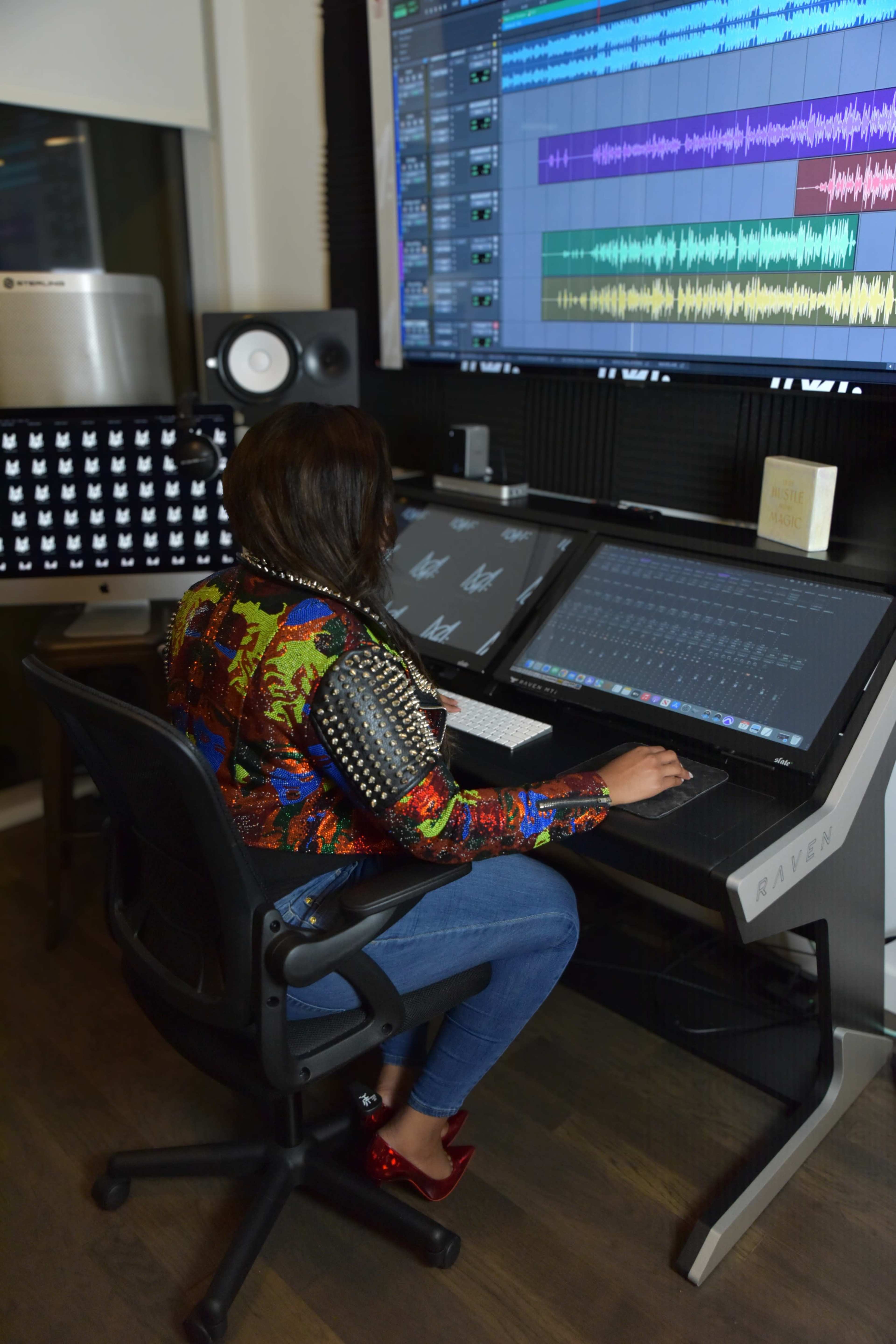 A person is sitting at a workstation with dual monitors displaying audio editing software and a digital audio workstation setup.