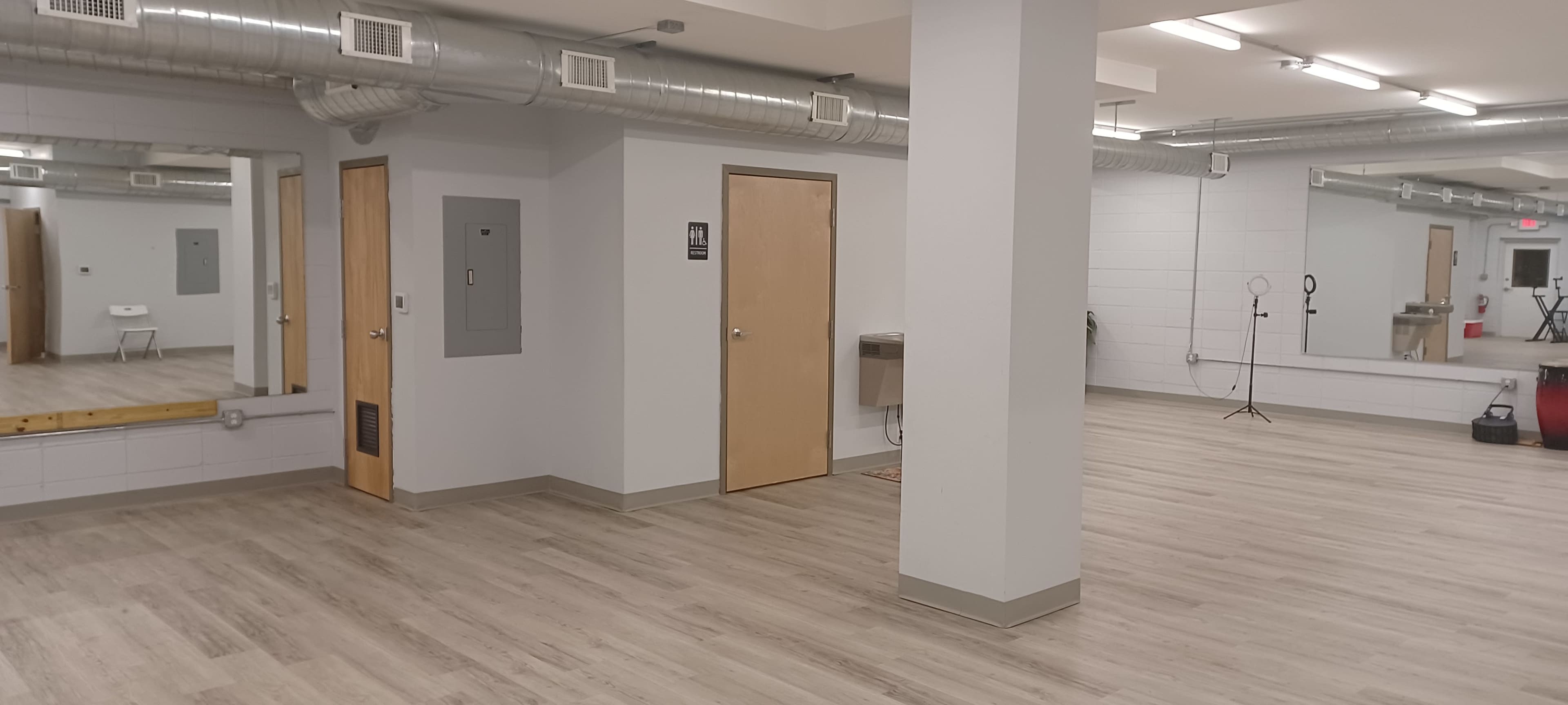The image shows a spacious, empty room with light wood flooring, featuring plain walls, a large mirror, a couple of doors, and exposed ductwork on the ceiling.