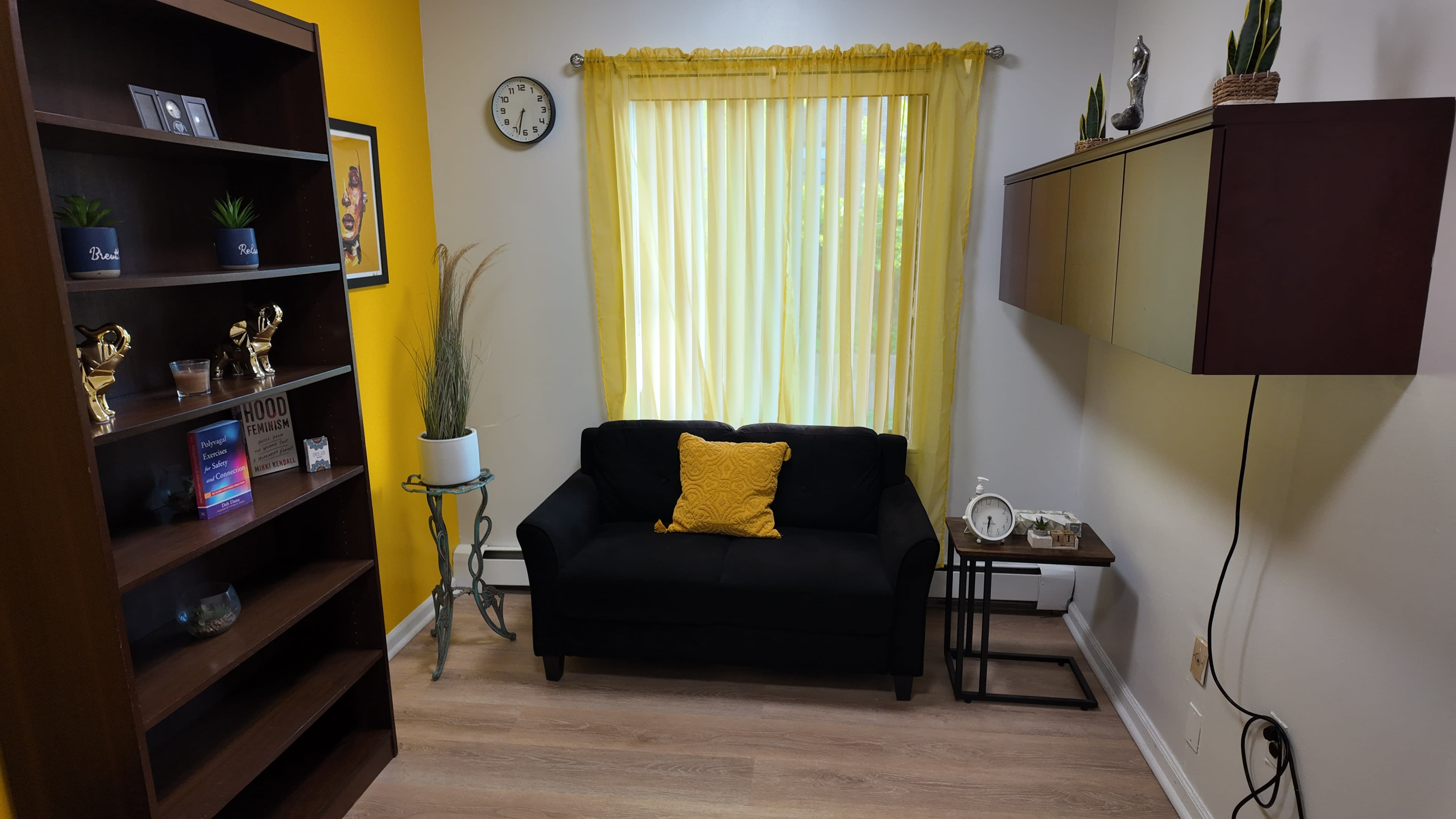 The image shows a small, cozy room with a black couch, a bookshelf with decor items, and a window with yellow curtains that let in light.