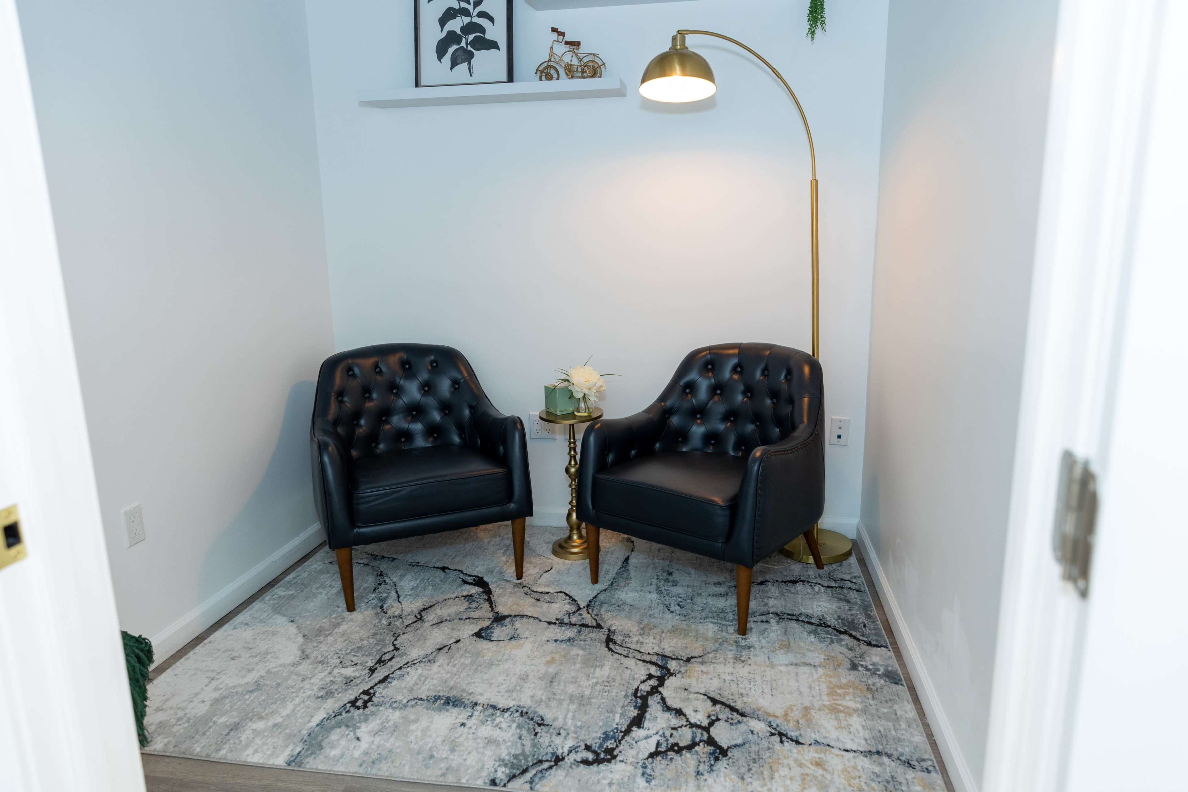 The image shows a small, minimalist corner of a room featuring two black upholstered chairs, a gold floor lamp, and a side table with a decorative arrangement on a patterned rug.