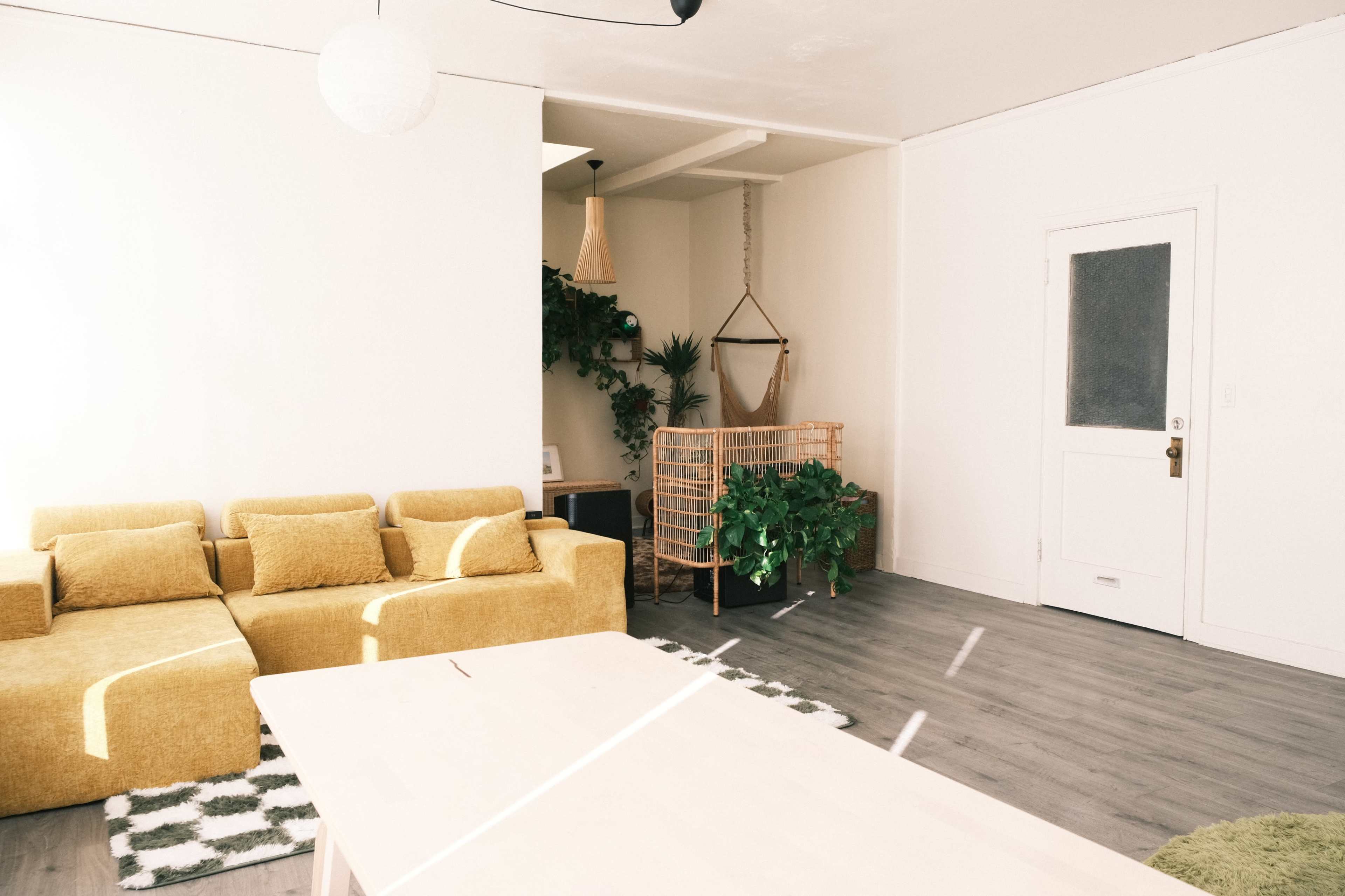 The image shows a minimalistic interior with a yellow sofa, a wooden dining table, and plants against a neutral-colored wall.