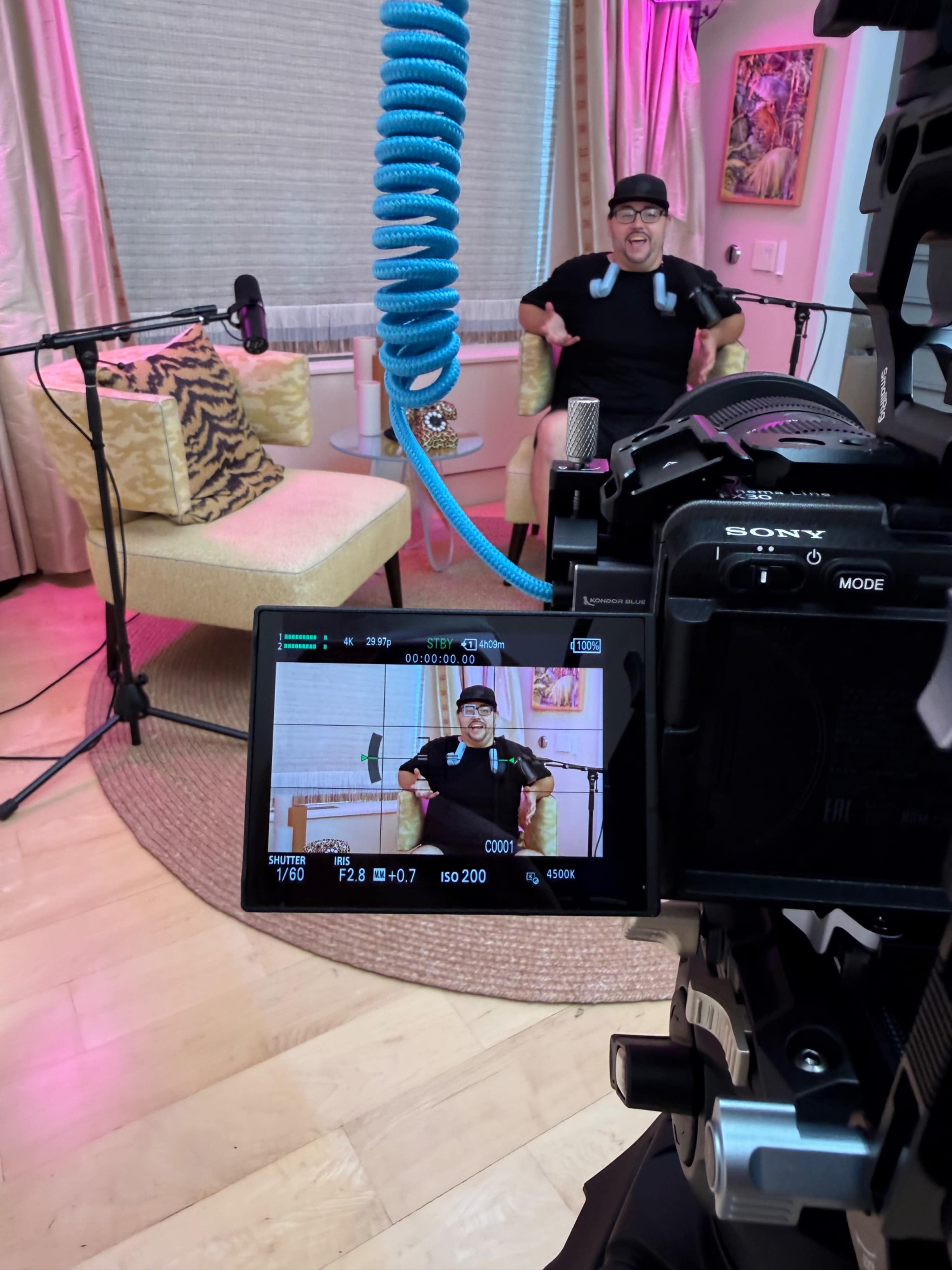 A man is seated in a yellow armchair in a brightly lit room, with a camera focused on him and microphones positioned nearby.