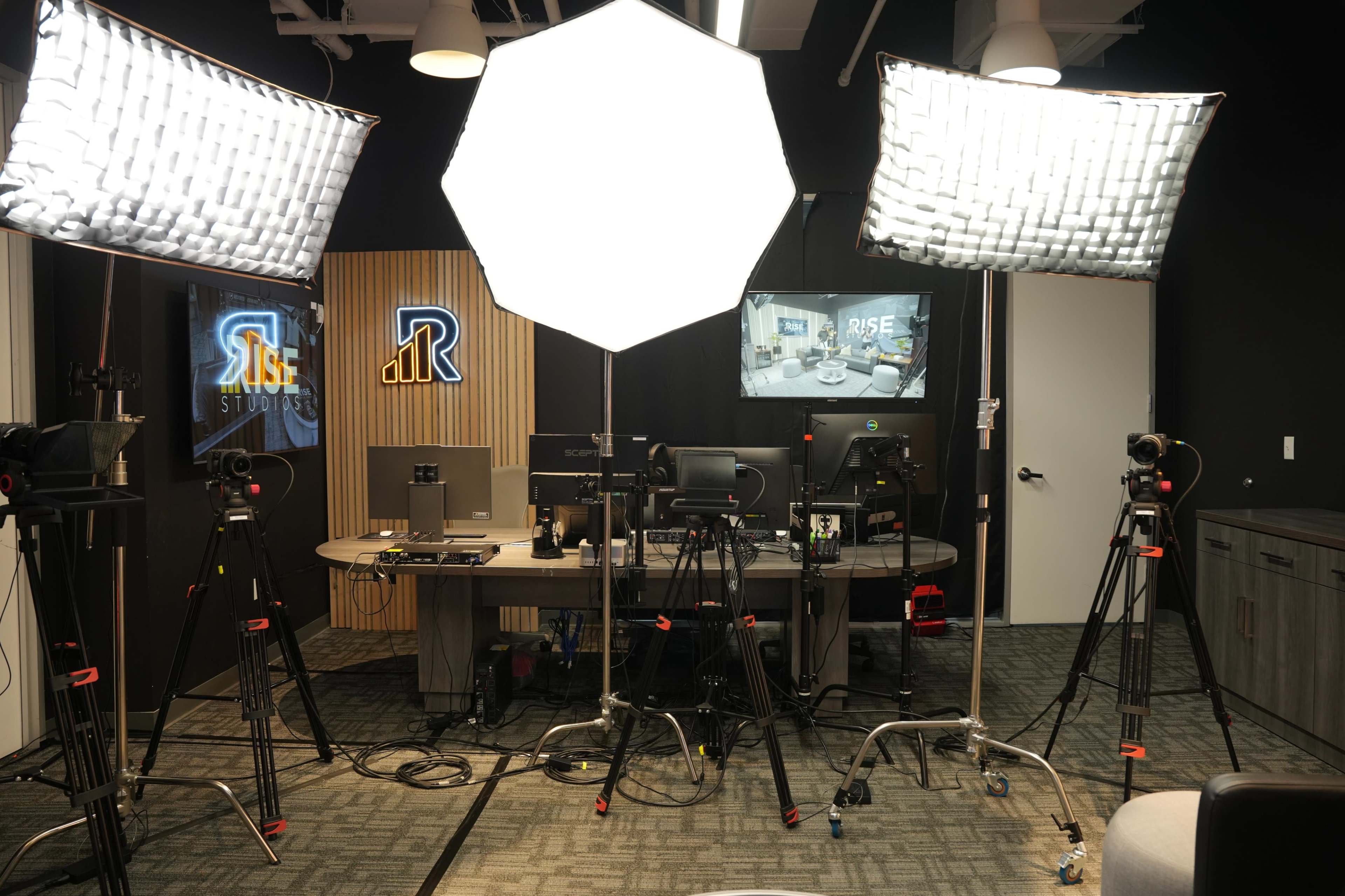 The image shows a production studio setup with multiple softbox lights, cameras on tripods, and a desk equipped with monitors and computers.