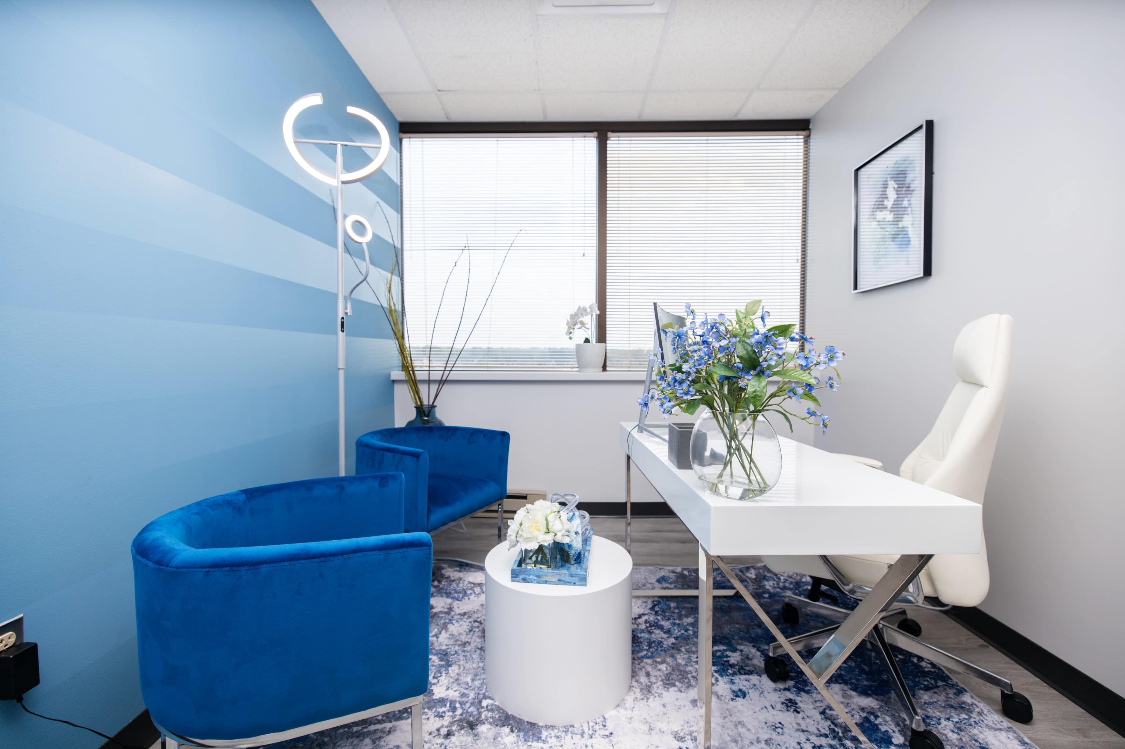 A modern office space features blue accent chairs, a white desk with a vase of flowers, and large windows letting in natural light.