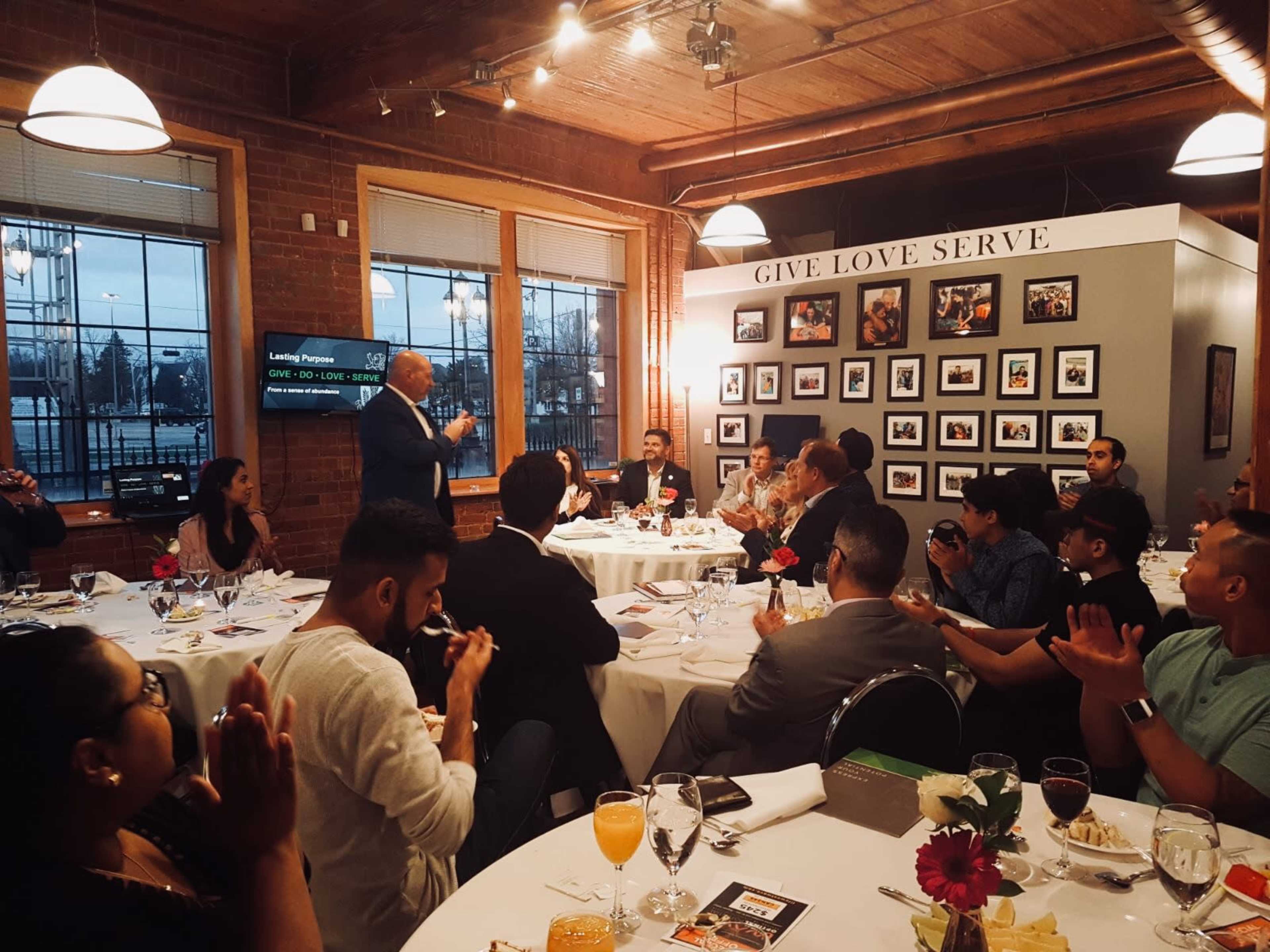 A speaker stands before a seated audience at a dinner event, with tables set and participants engaged in applause.