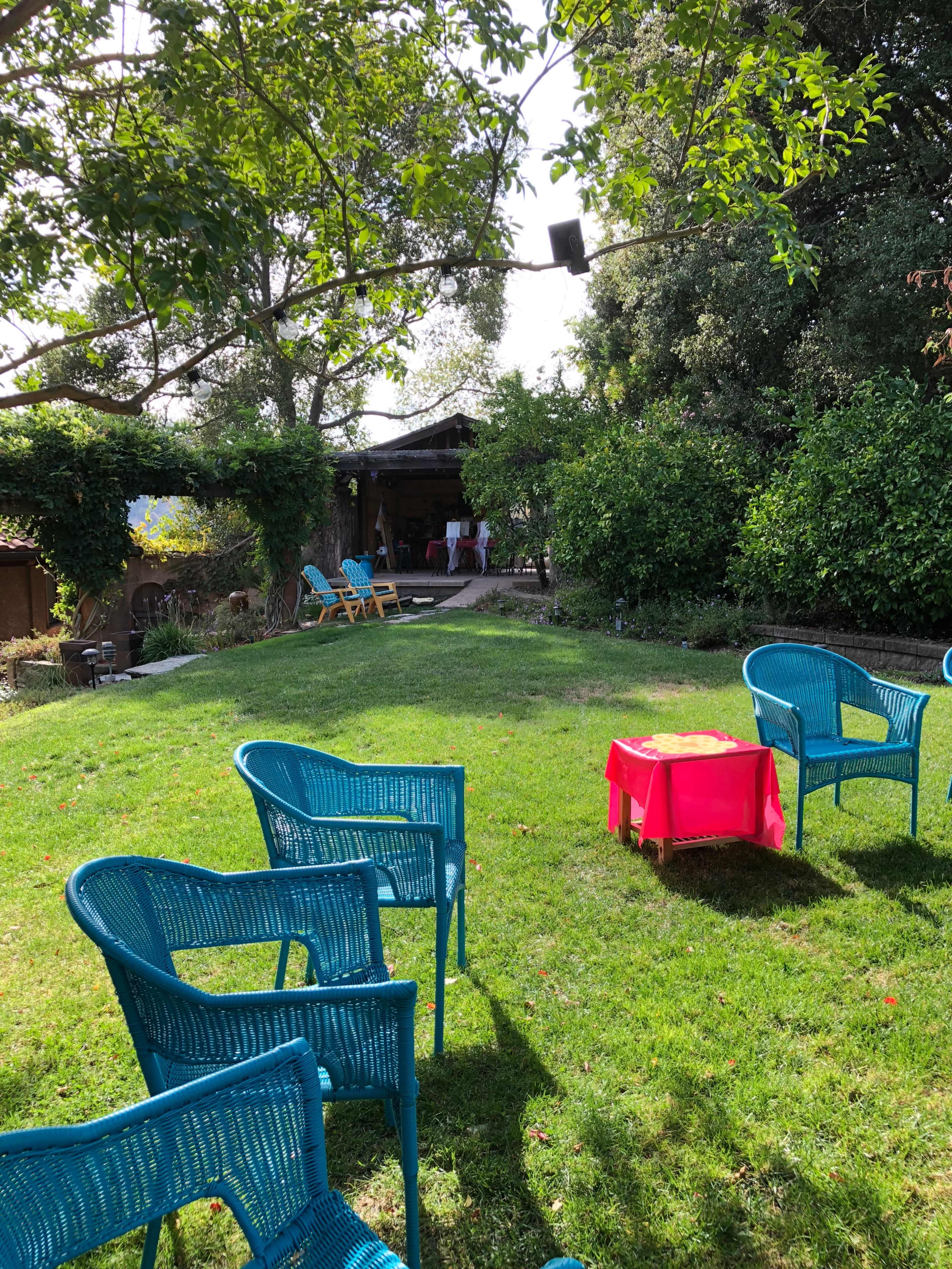 A grassy yard features blue wicker chairs arranged around a small red table, with a rustic structure and greenery visible in the background.