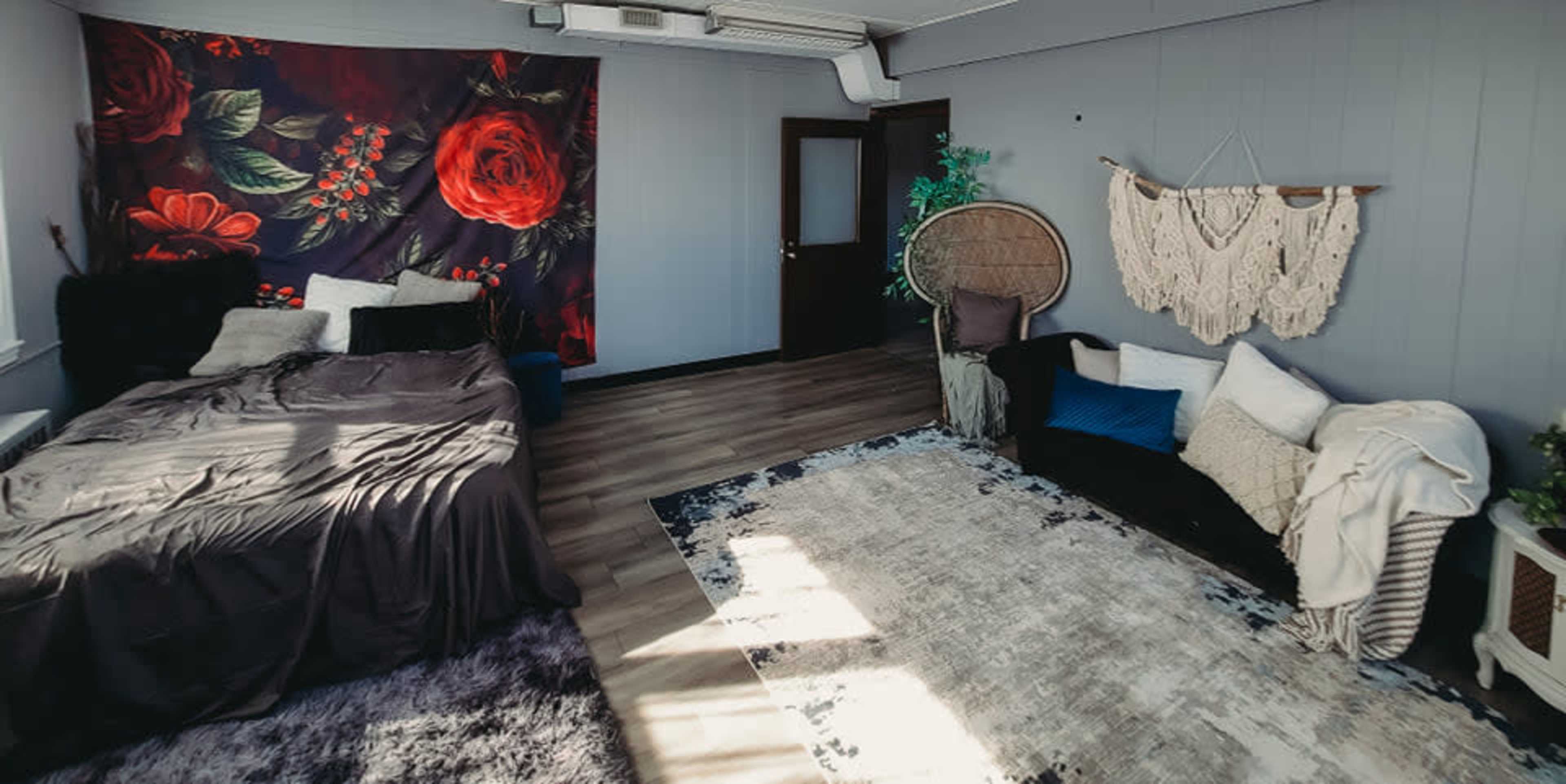 A room with a bed covered in dark bedding, a large floral tapestry on one wall, and a cozy seating area with a rug and decorative throw pillows.