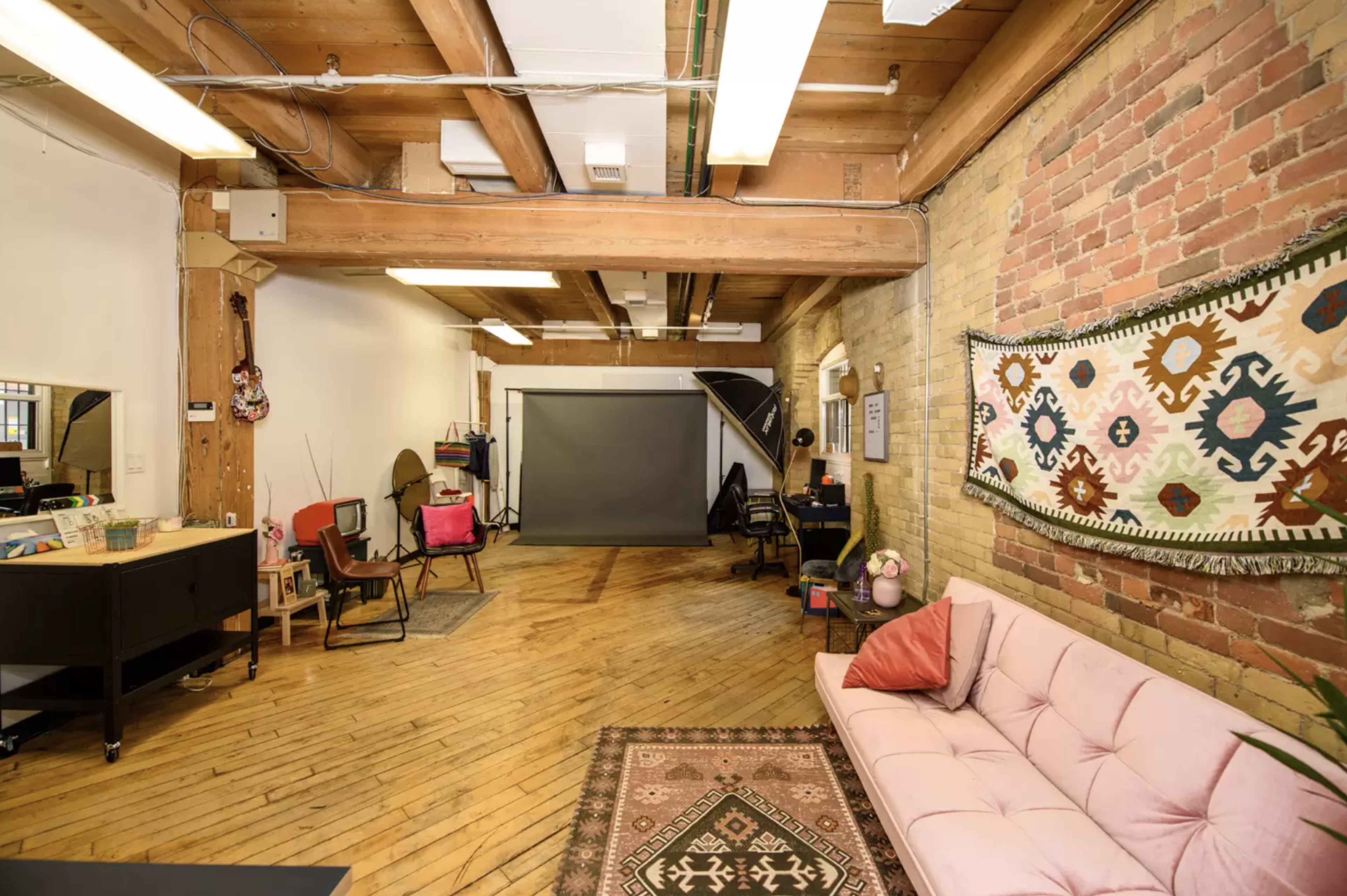 A spacious studio with wooden beams, brick walls, and a pink couch, featuring a backdrop for photography and various furniture arranged around the room.