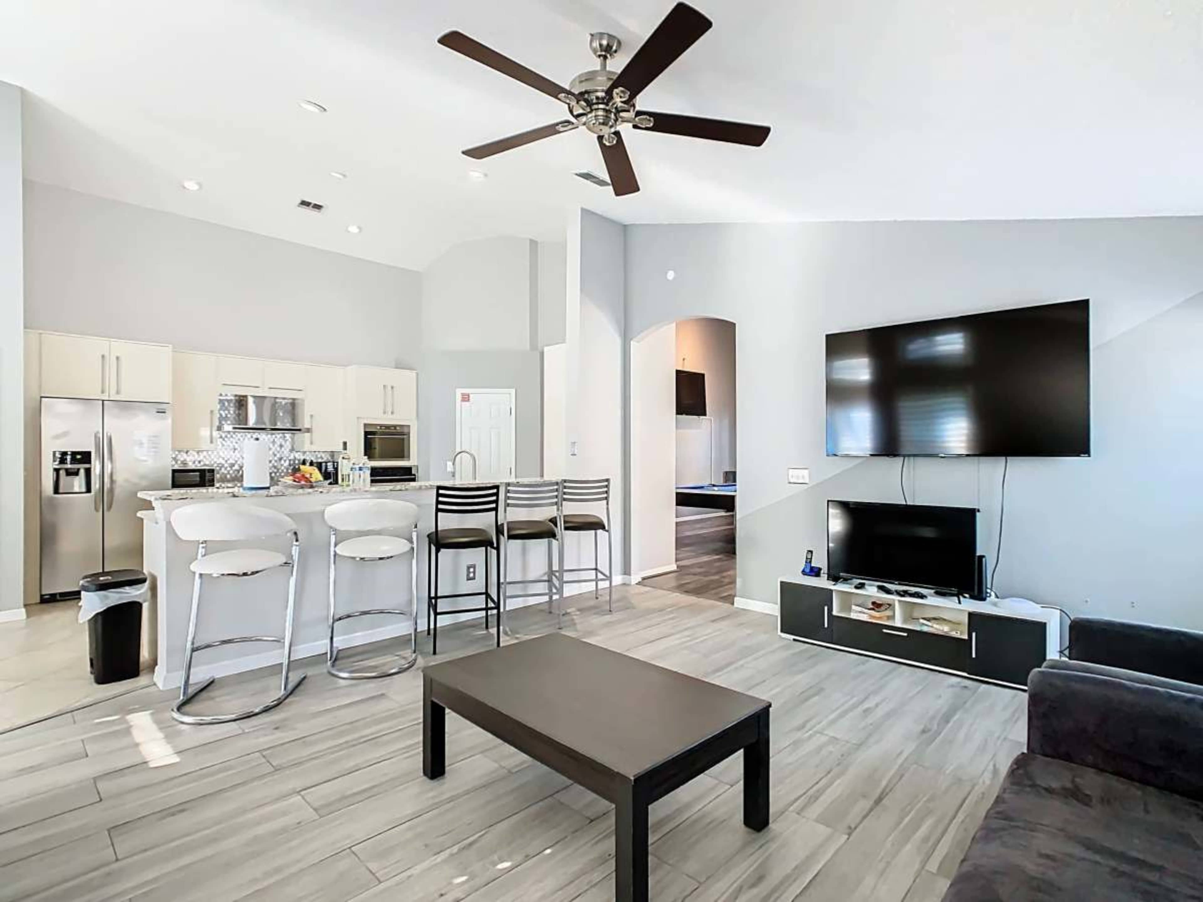 The image shows a modern living room that connects to a kitchen, featuring a large television mounted on the wall, a black coffee table, and bar stools at the kitchen counter.
