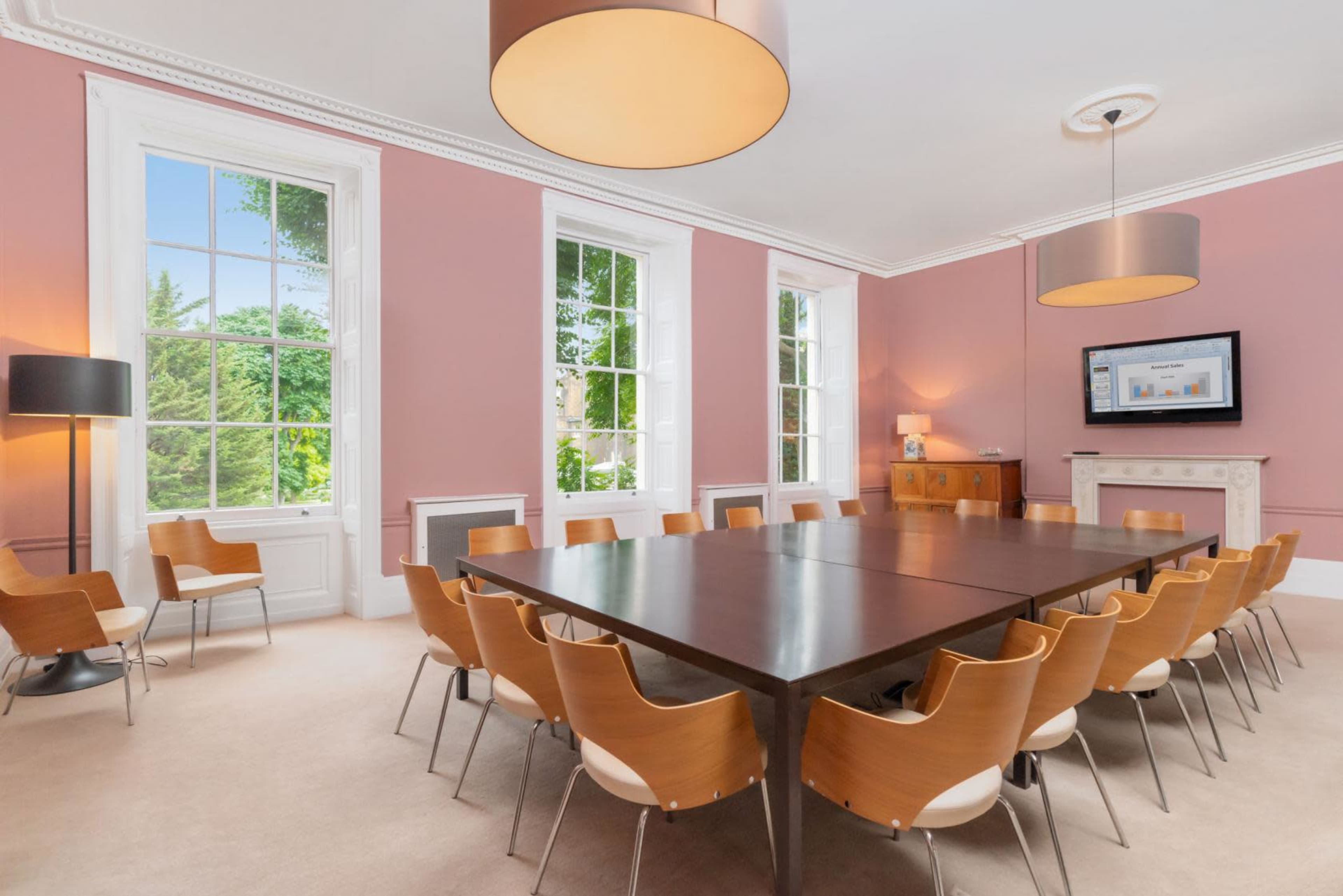 A spacious meeting room with a large rectangular table surrounded by wooden chairs, large windows, and a television mounted on the wall.